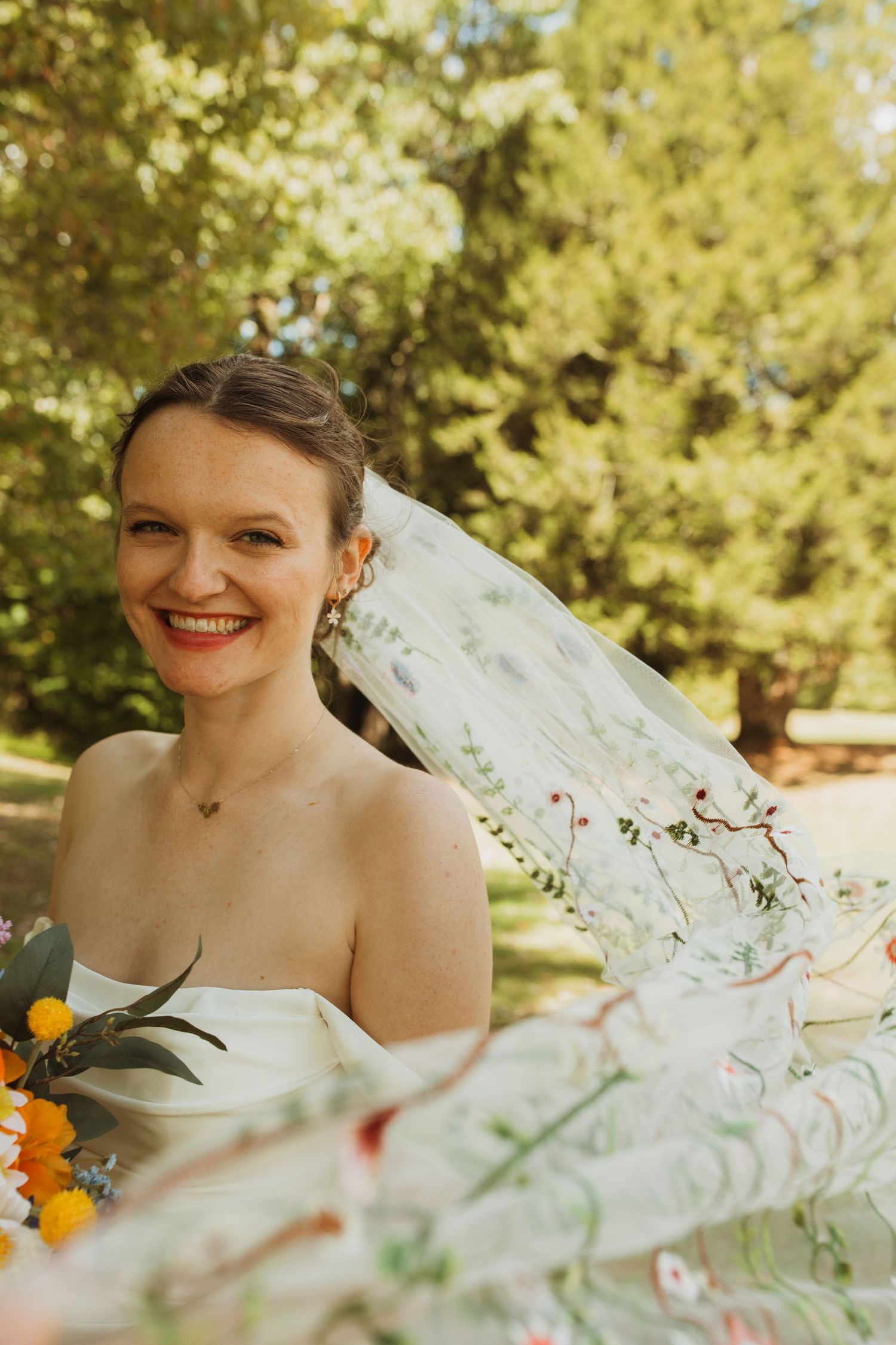 A radiant bride wearing a strapless dress and veil poses outdoors with bright flowers.