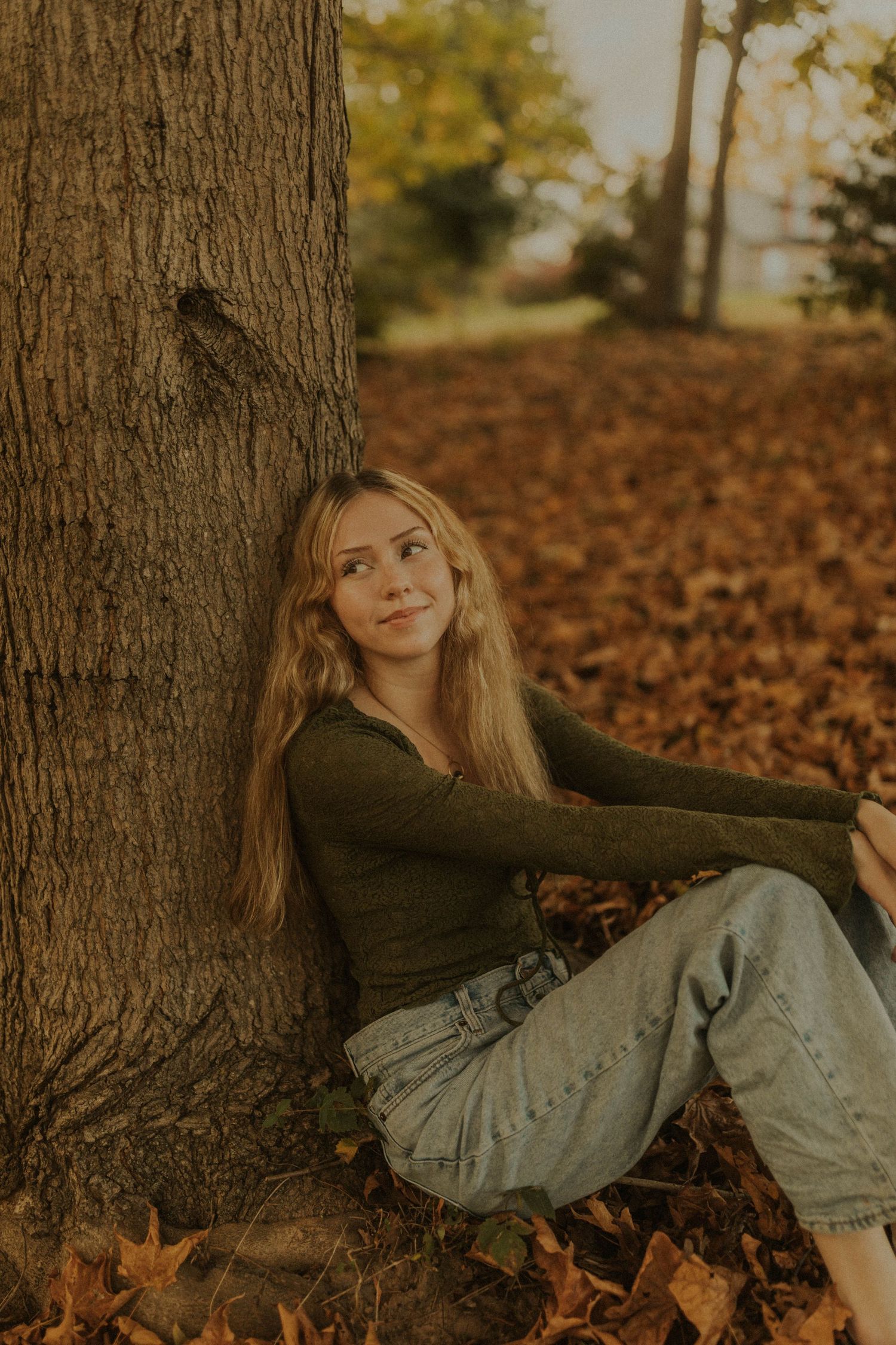 Person in green sweater sitting against autumn tree trunk surrounded by fallen leaves.