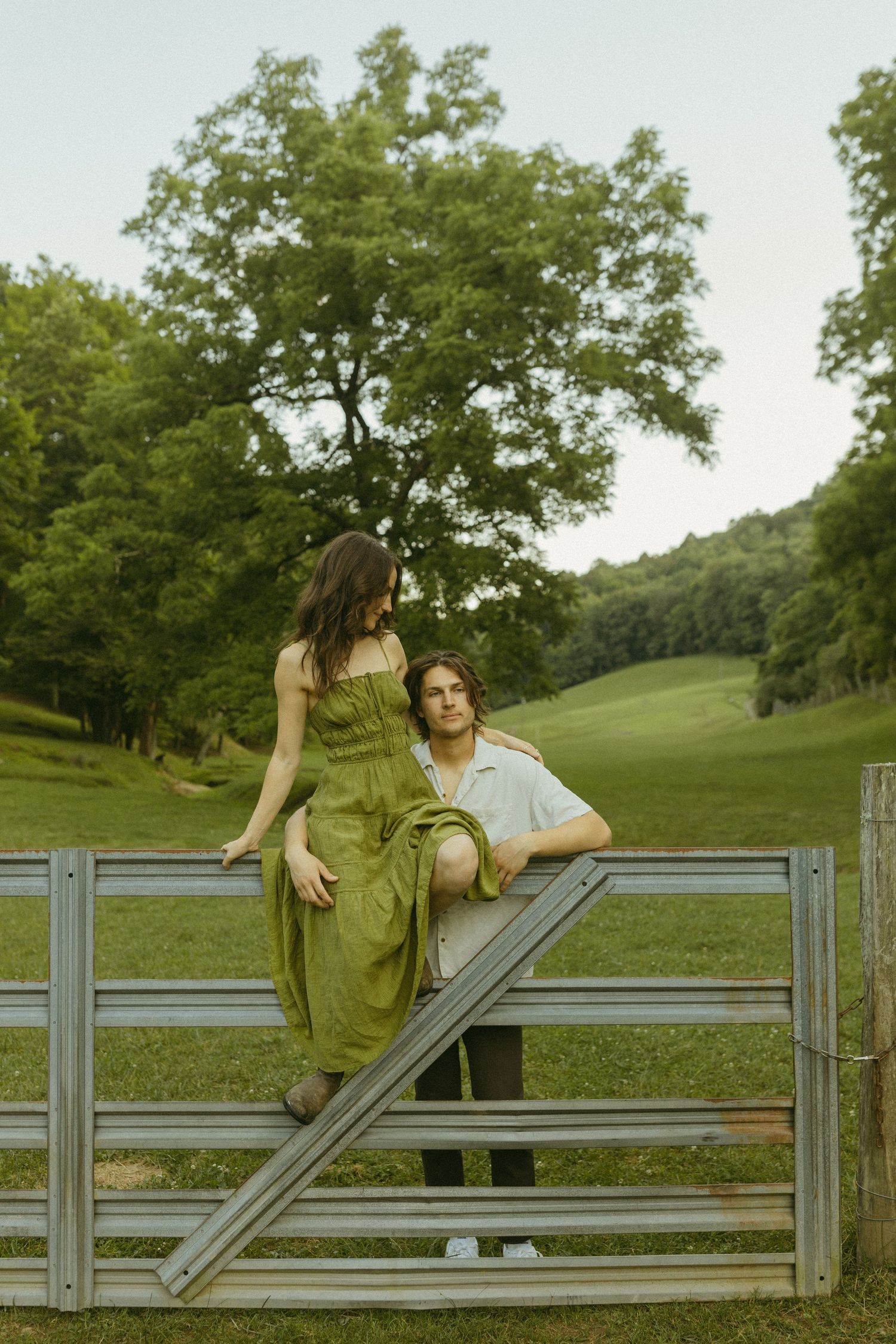 Couple shares a quiet moment while perched on a metal farm gate in a green field.