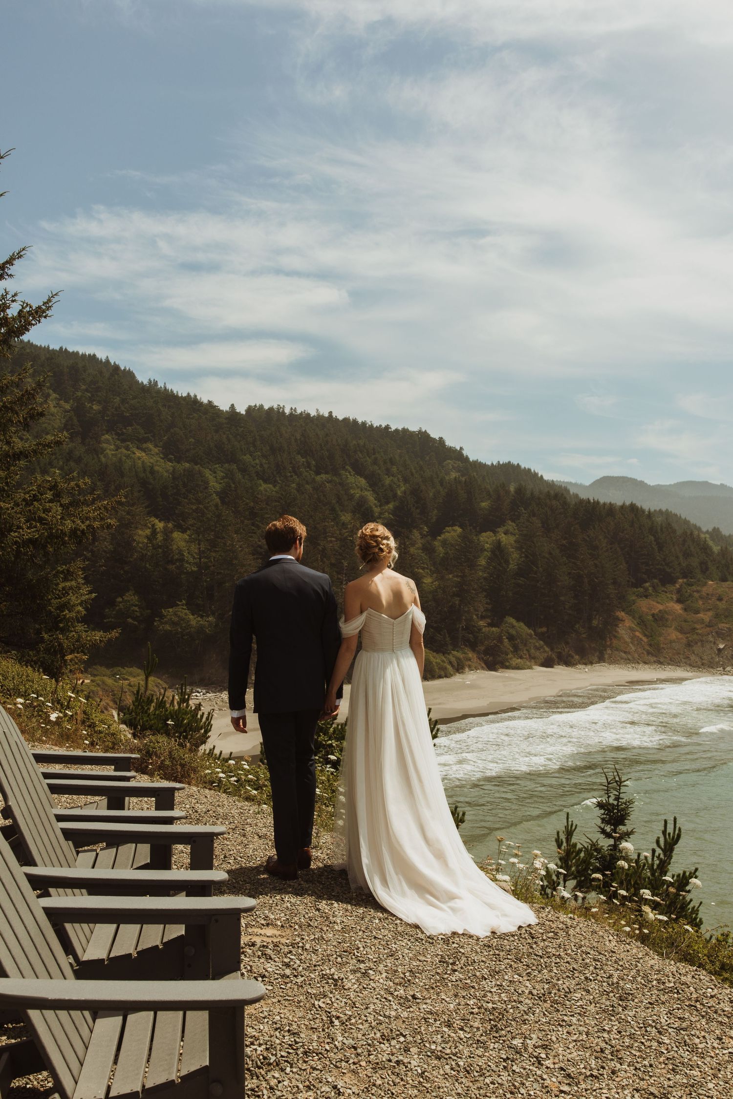 A couple in wedding attire stands on viewing deck overlooking a scenic coastal beach.