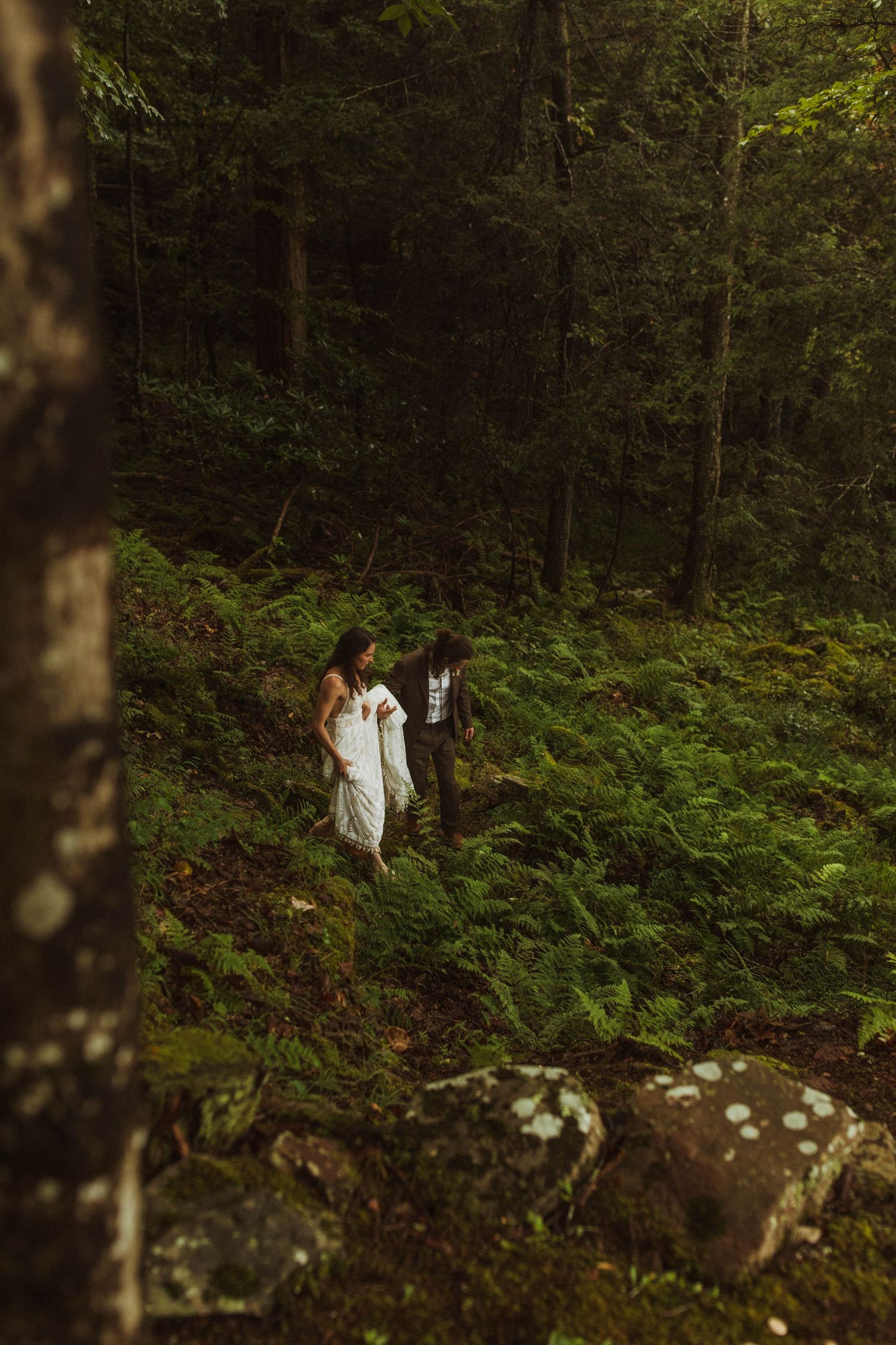 Couple walking through fern-covered forest floor in wedding attire.