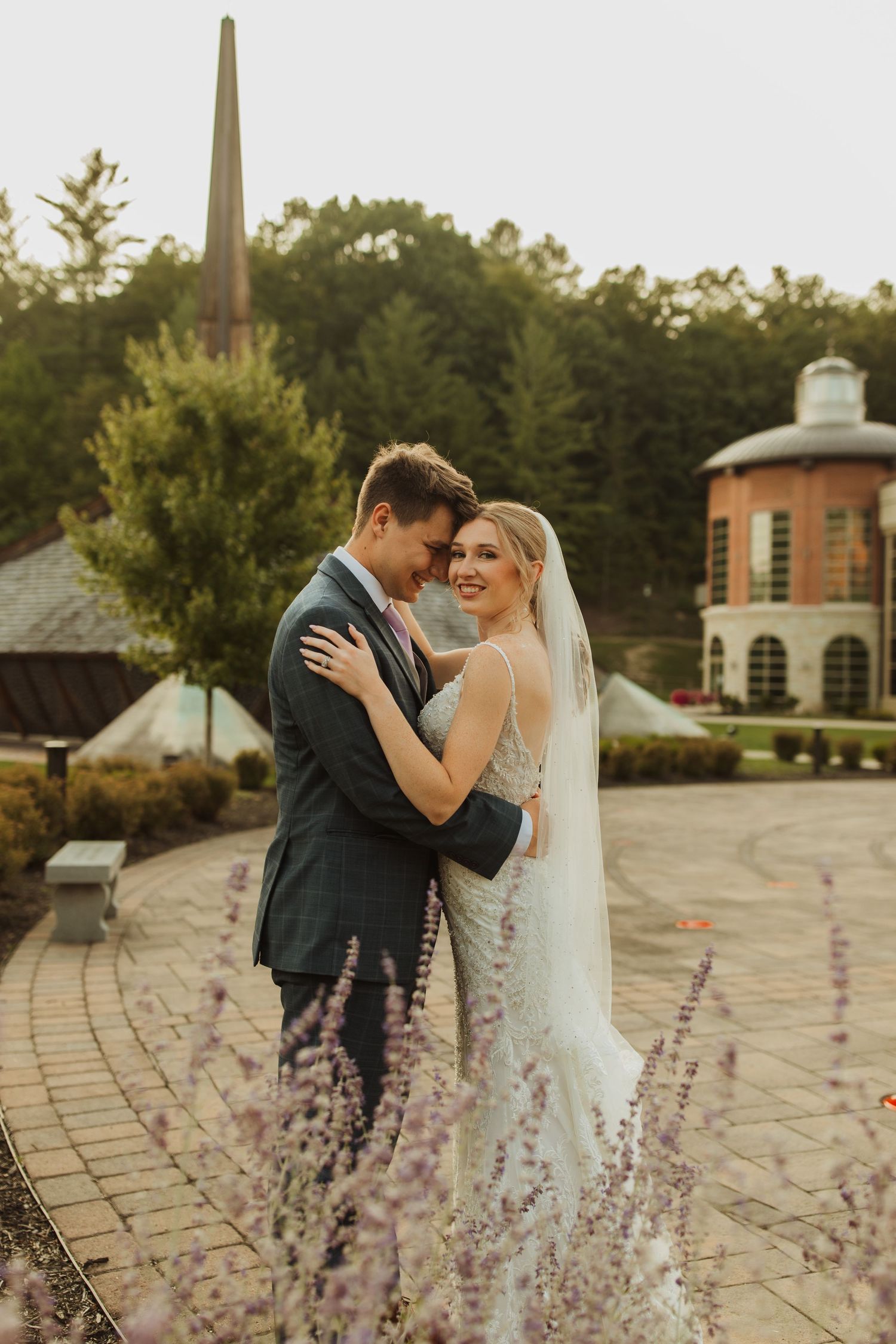 Romantic wedding couple embraces on brick pathway with lavender flowers and church spire in background at sunset.