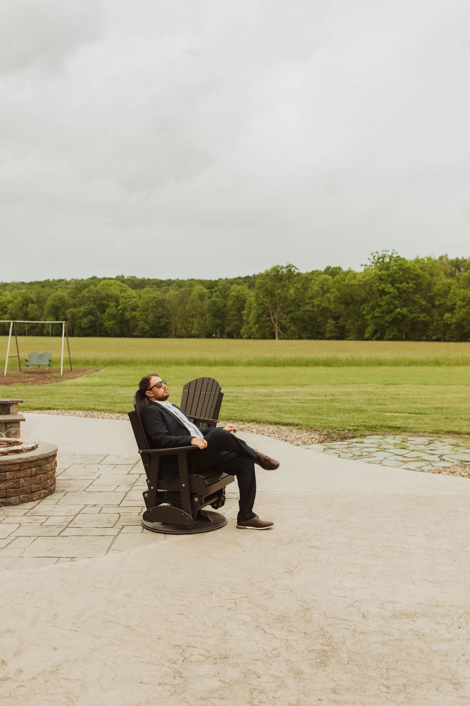 A person relaxes in an Adirondack chair on a patio overlooking a grassy field with soccer goals.