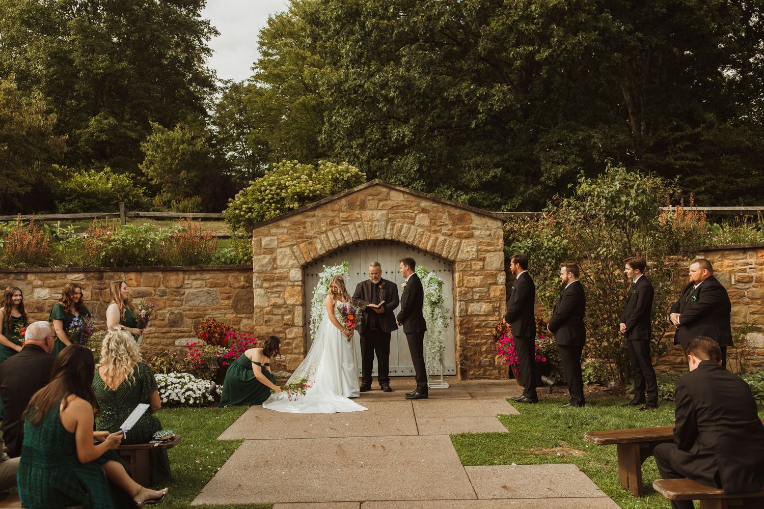 Outdoor wedding ceremony taking place under a rustic stone archway with guests seated on benches.