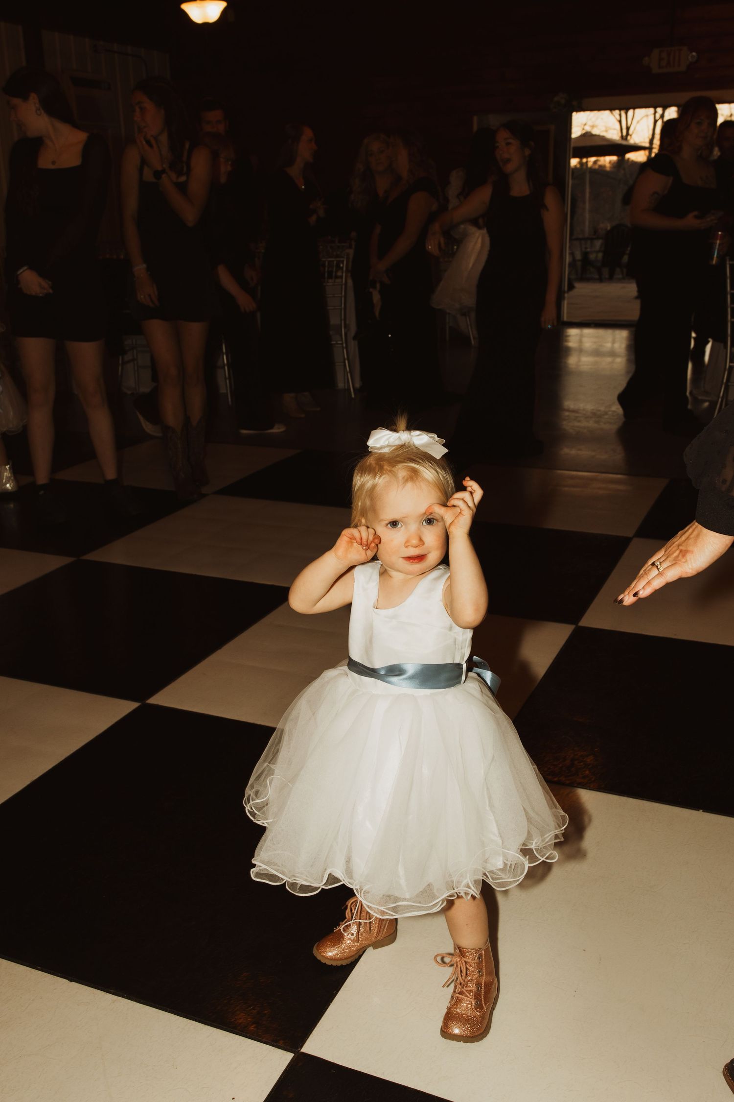 Young flower girl in white tulle dress dancing on checkered dance floor at wedding reception.