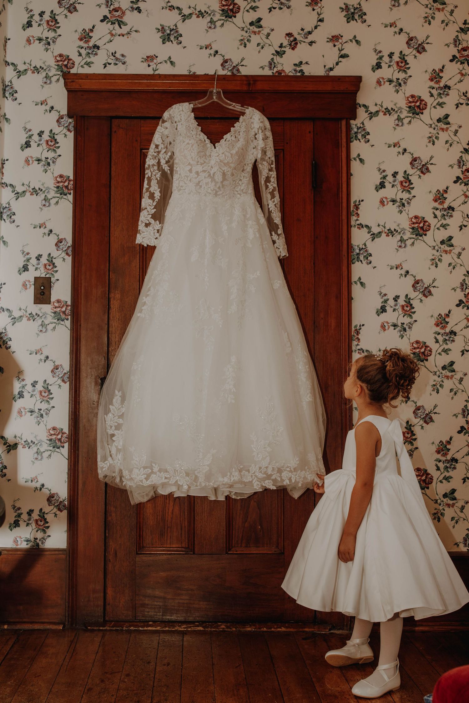 An elegant lace wedding dress hangs on display with a young attendant in white nearby.