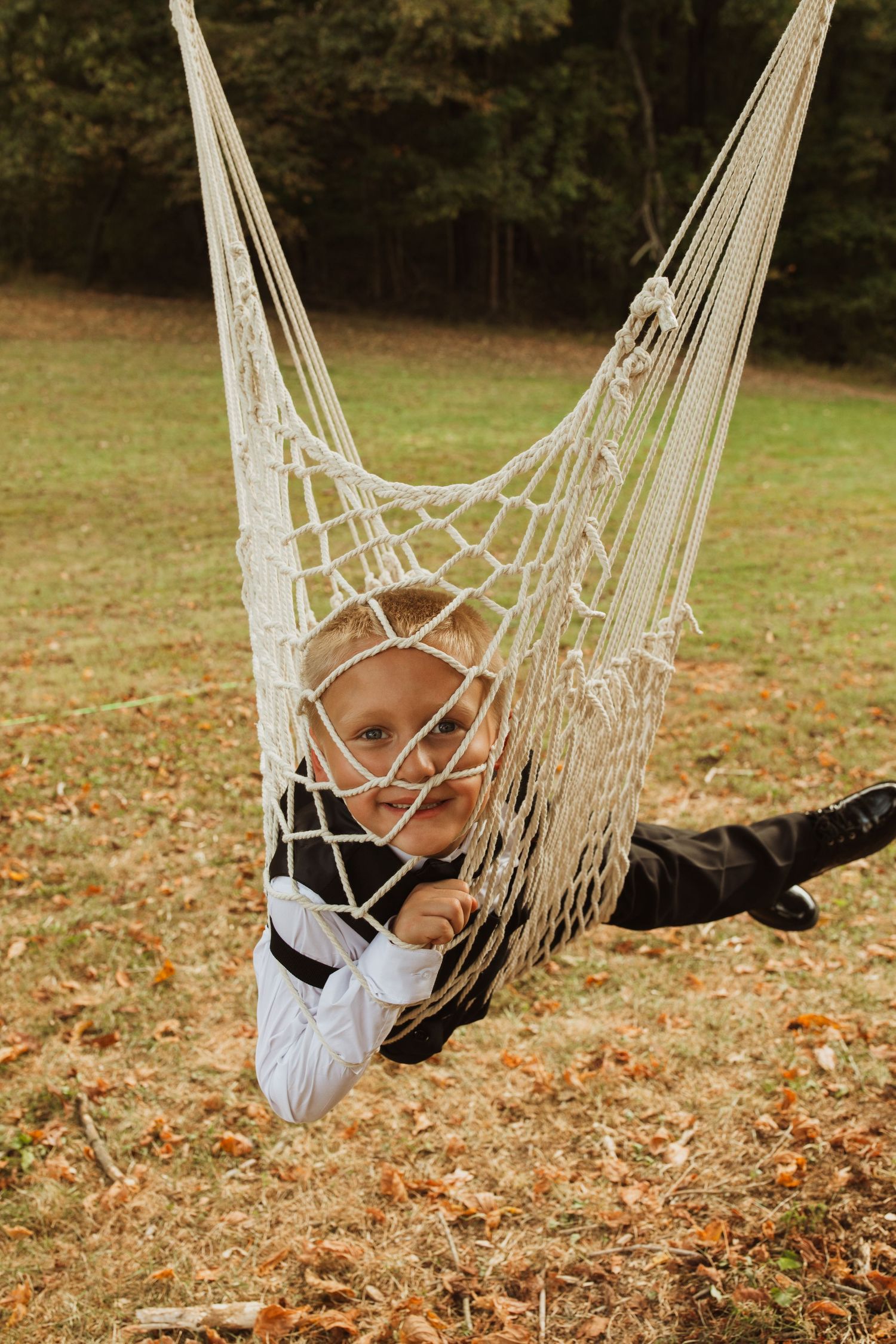 A person in formal attire lounges playfully in a macrame hammock surrounded by fallen autumn leaves.