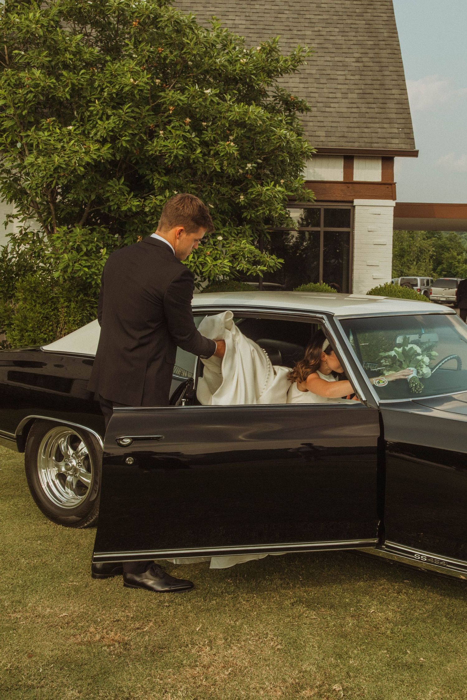 A person in formal attire helps with a car door against a Tudor architecture backdrop.