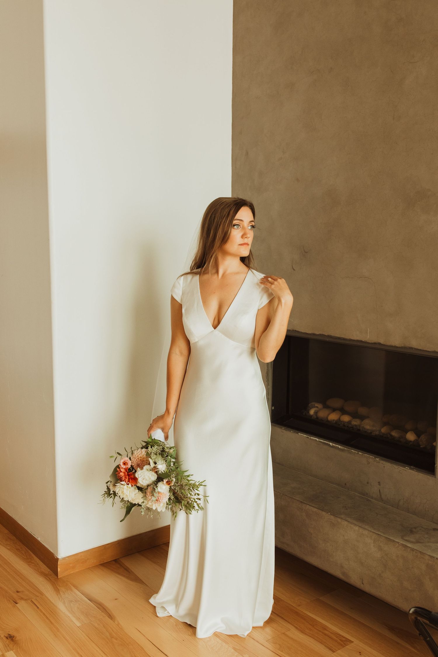 Elegant bride in white v-neck dress holding bouquet by modern fireplace.