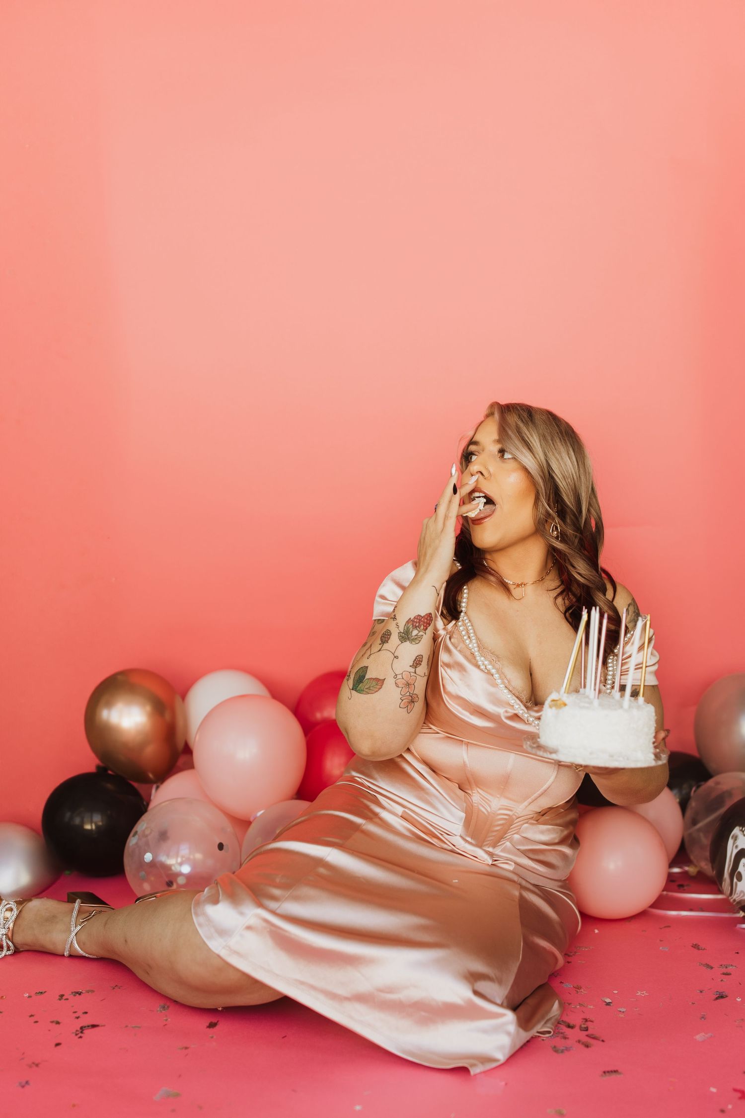 Woman in pink silk dress laughing with birthday cake surrounded by balloons against coral background. birthday photoshoot