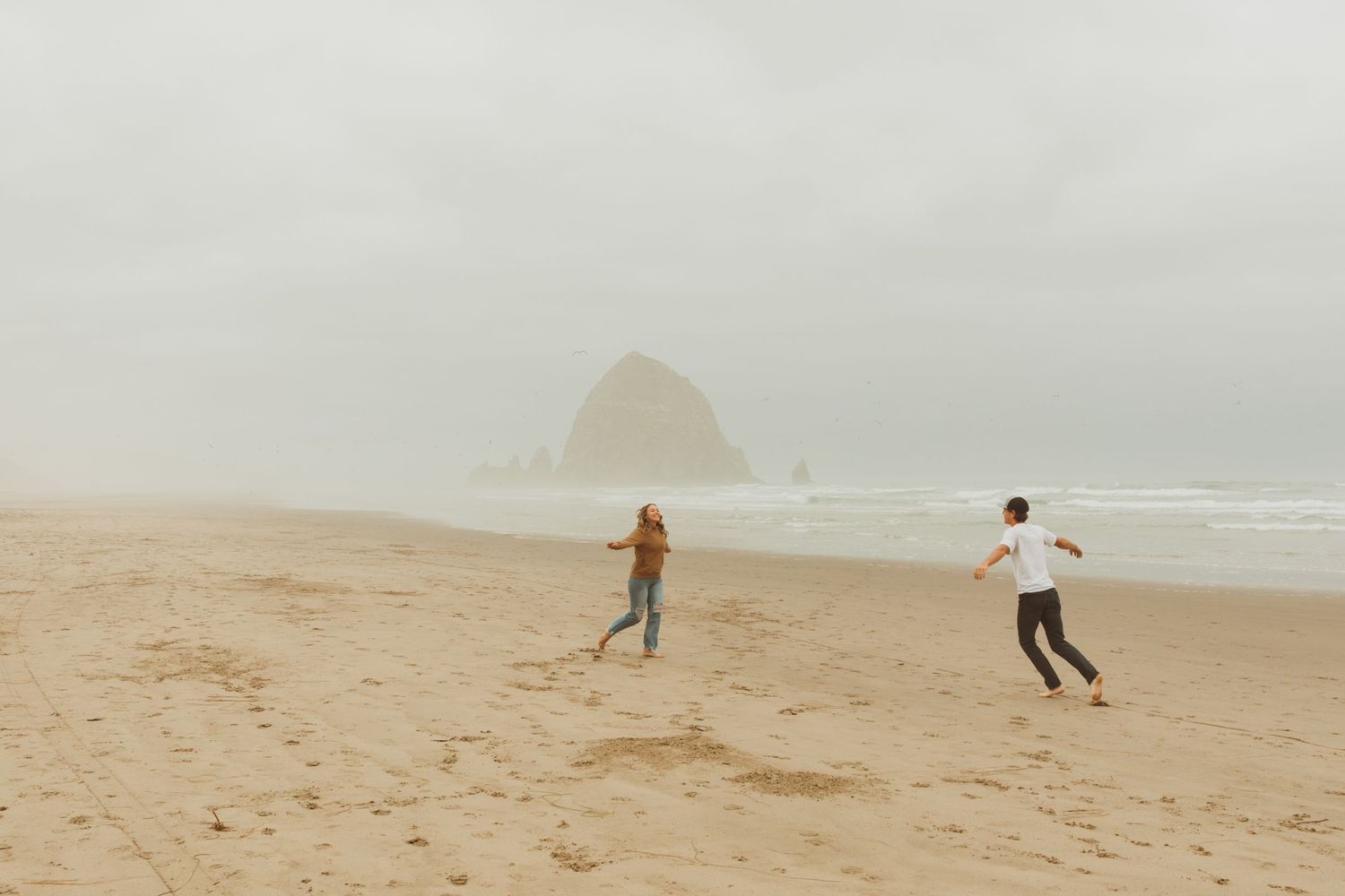 Two people running playfully on misty beach with Haystack Rock in background.