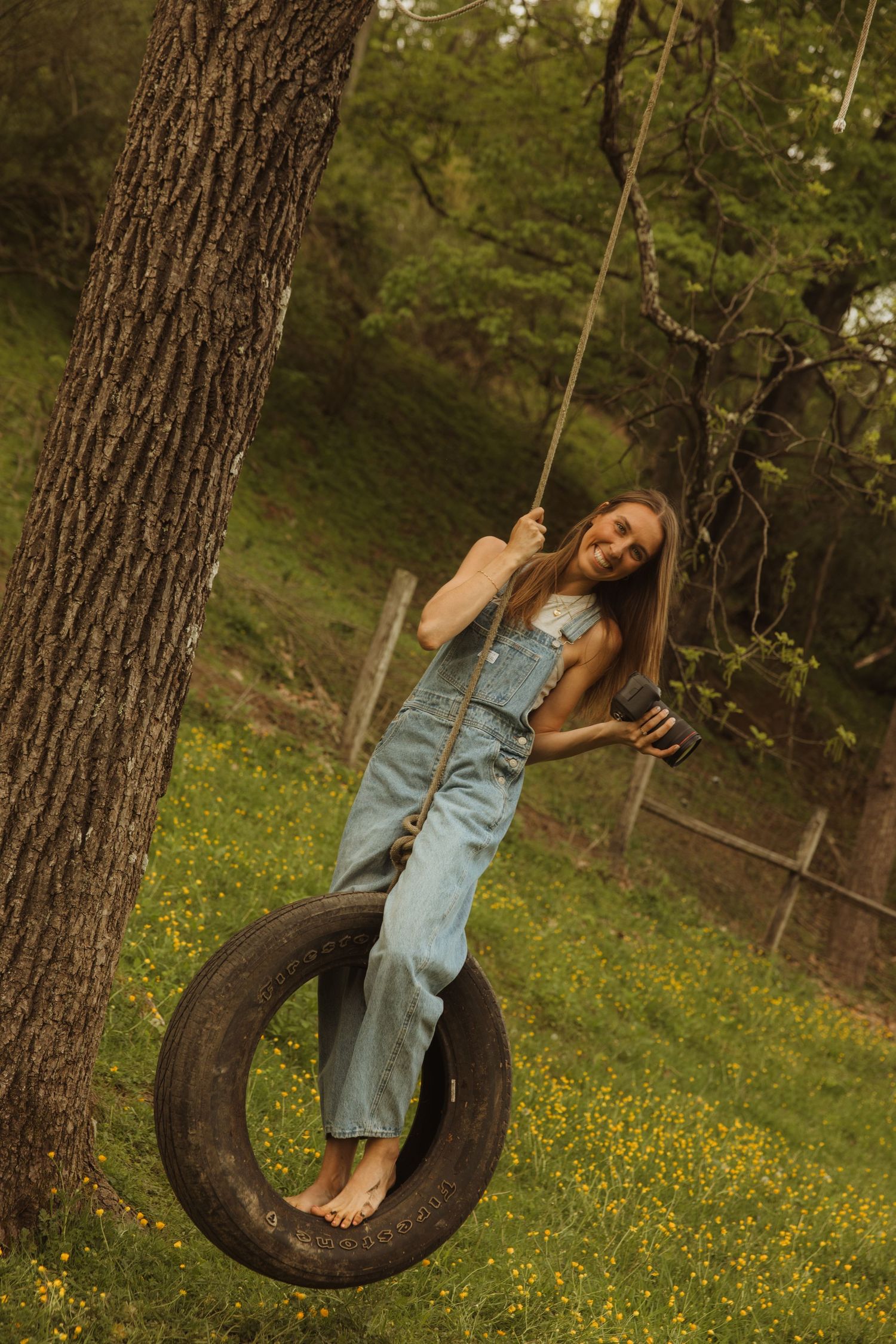 A vintage tire swing hangs from a large tree in a grassy yard with yellow wildflowers in bloom.