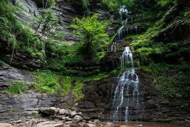 A multi-tiered waterfall flows down moss-covered rock face in a verdant forest setting.