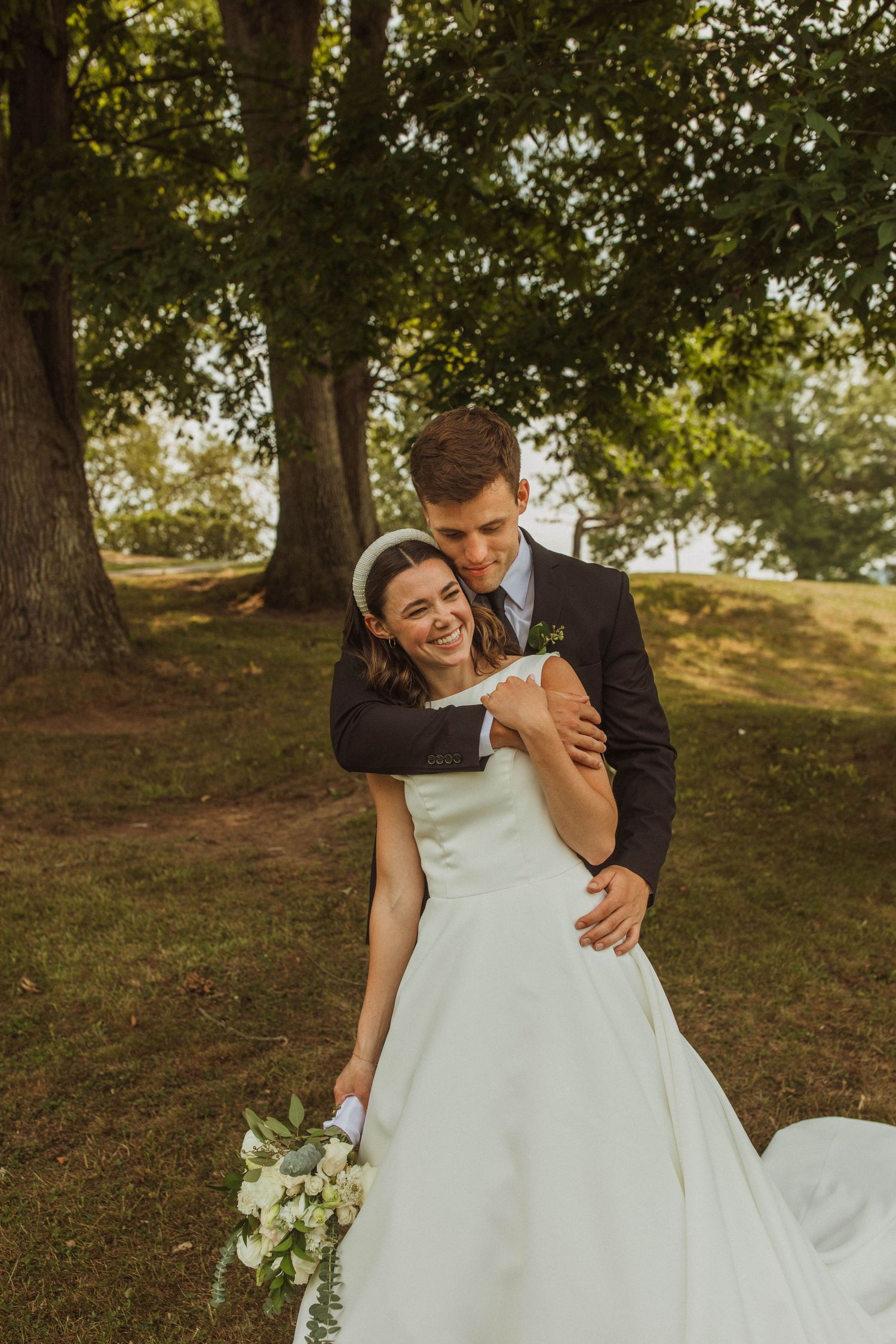 A romantic wedding portrait outdoors with the couple embracing under tall trees during golden hour.