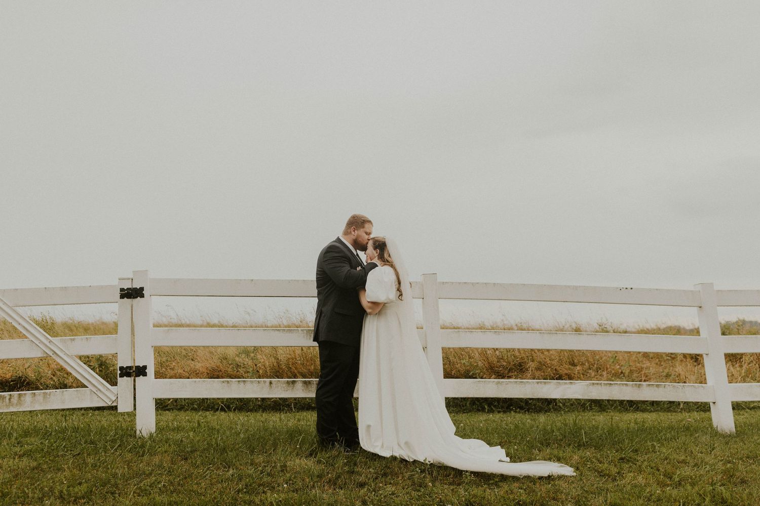 A romantic wedding moment against a white fence on an overcast day.