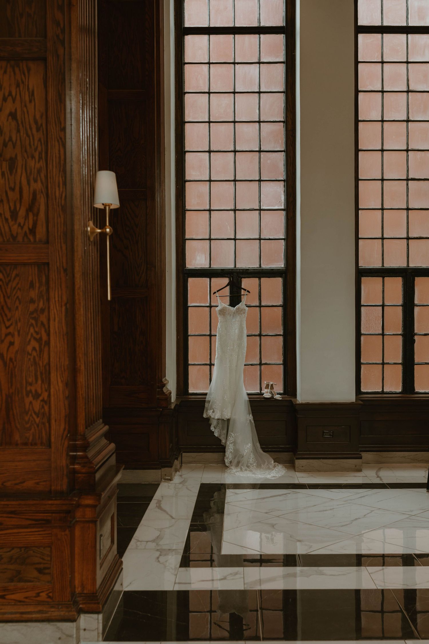 Elegant white wedding dress hangs in front of tall windows with wood paneling.