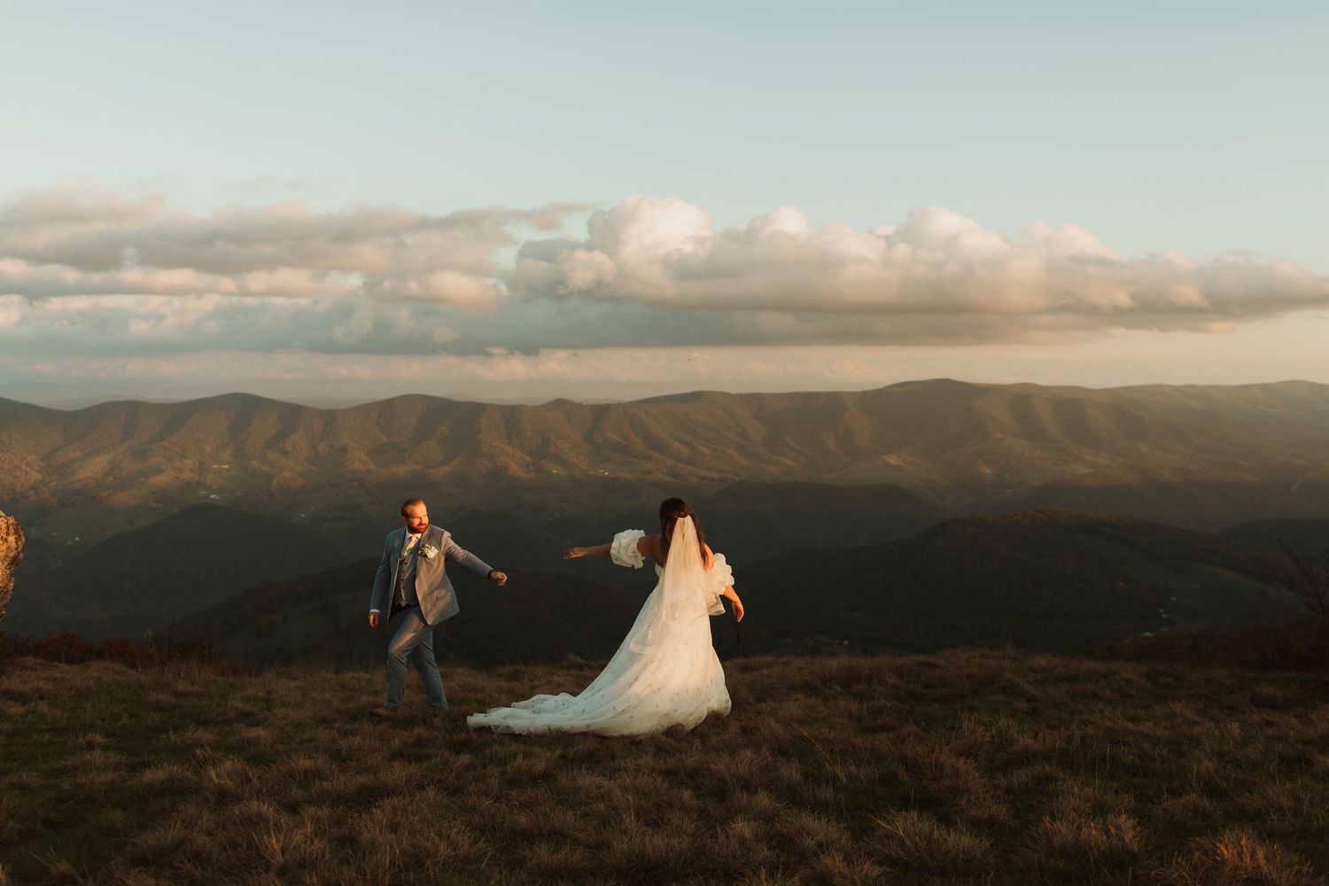 A couple walks along a mountaintop meadow with dramatic mountain ranges in the background.