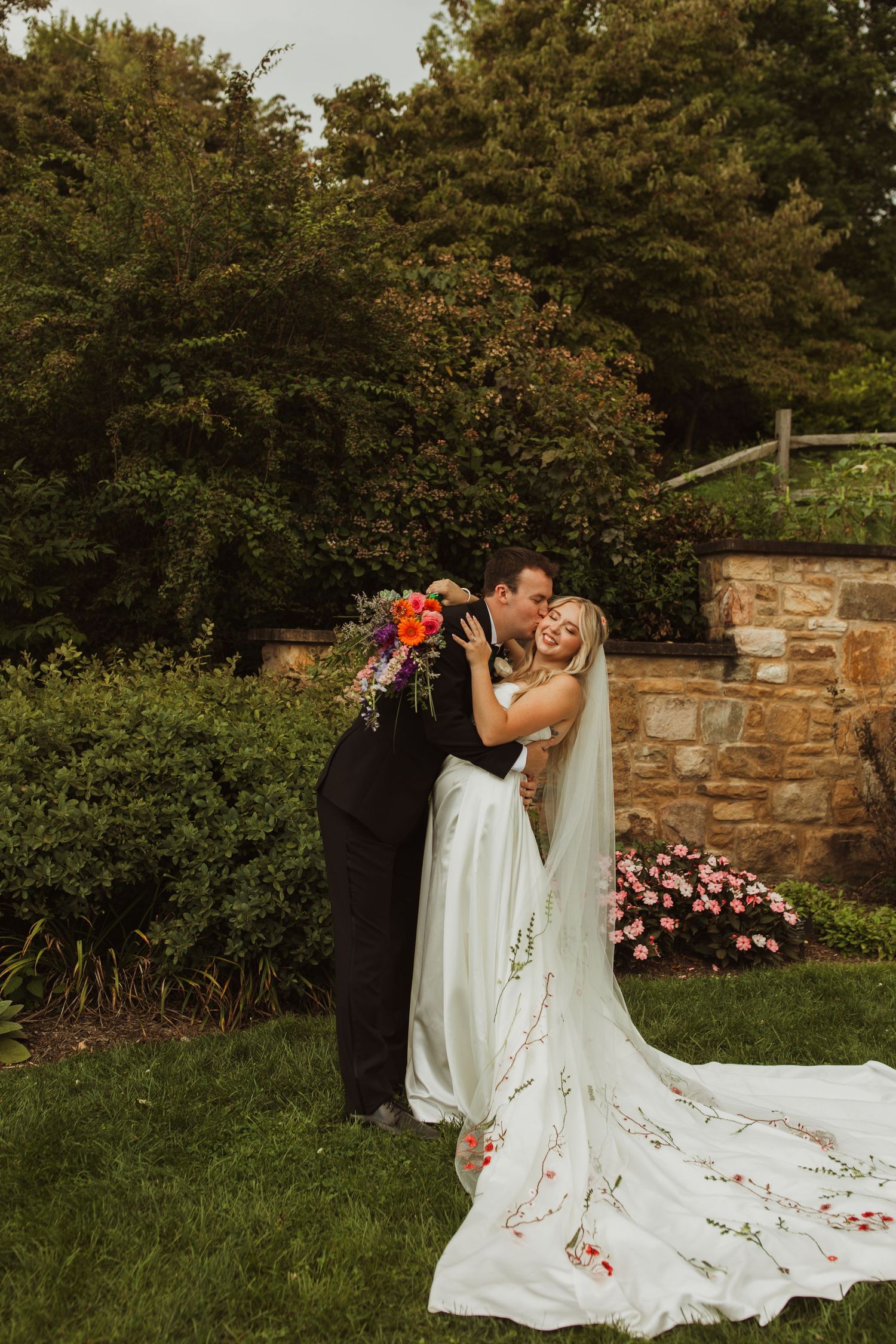 Newlyweds share a joyful moment in a garden with stone wall and colorful florals.