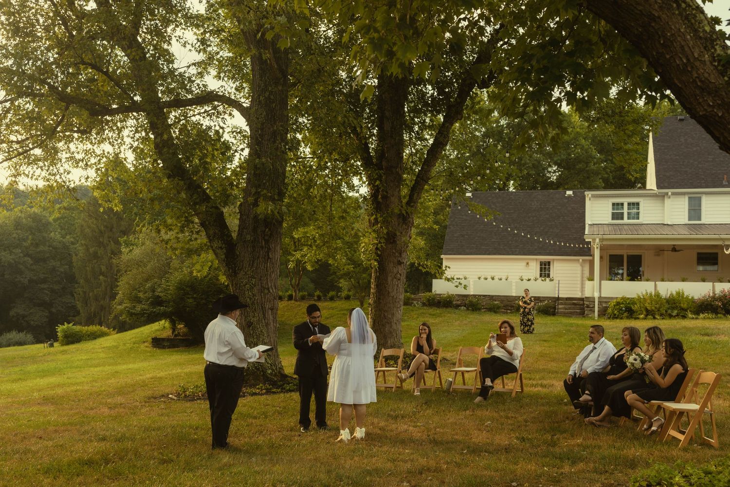 Intimate backyard wedding ceremony under trees with guests seated on wooden chairs.
