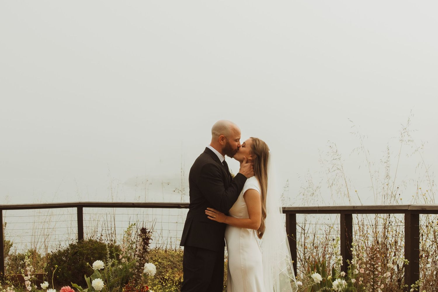 Newlyweds share a romantic kiss on a misty overlook surrounded by wildflowers and a rustic railing.