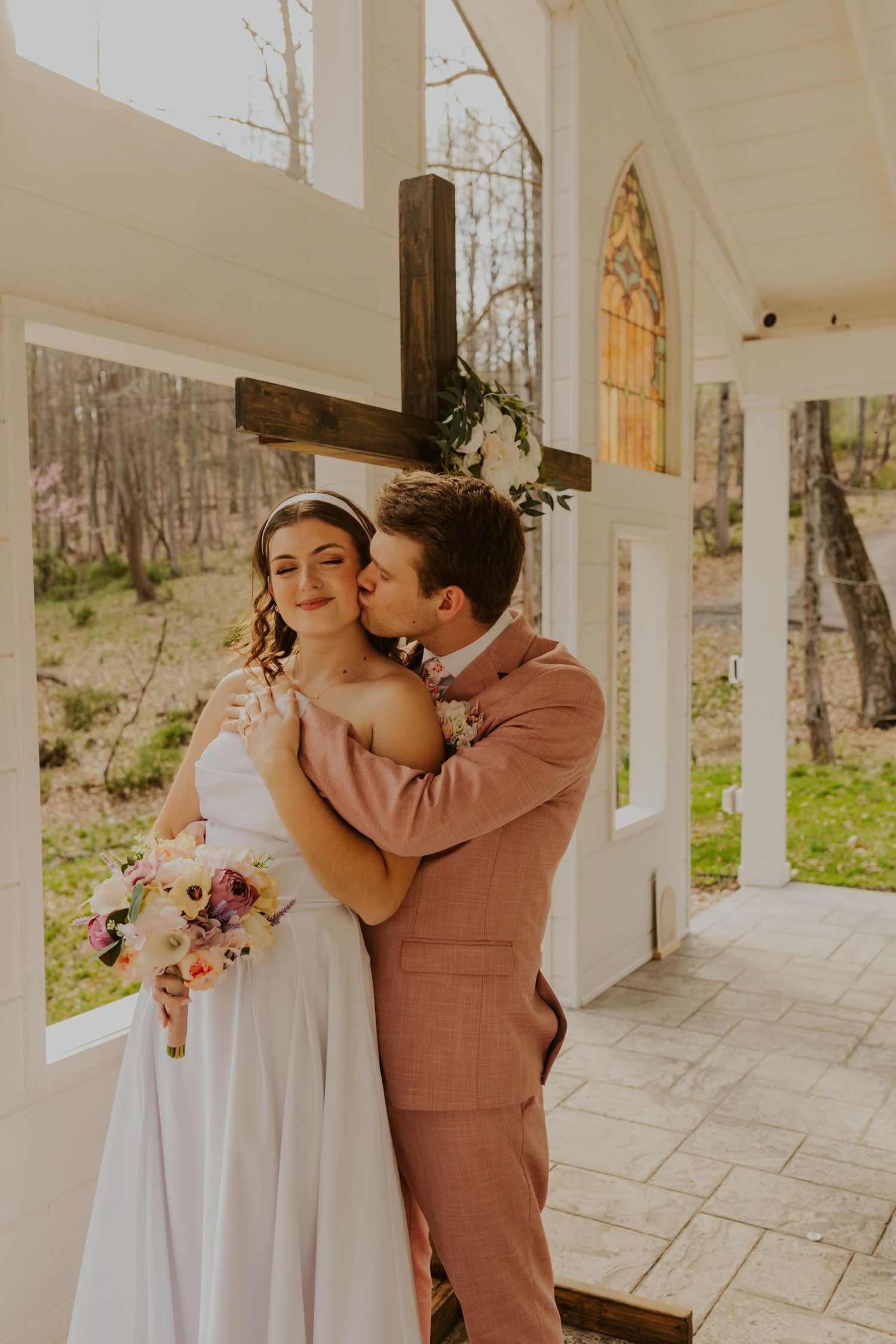 Romantic wedding portrait by wooden cross with couple in blush suit and white dress.