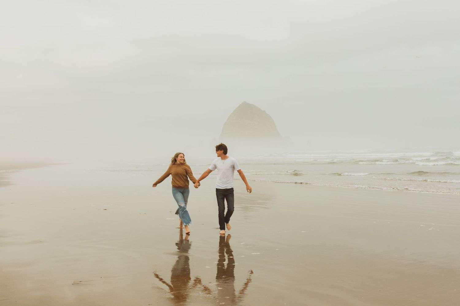 Couple walking hand in hand on wet sand reflecting misty Haystack Rock at beach.