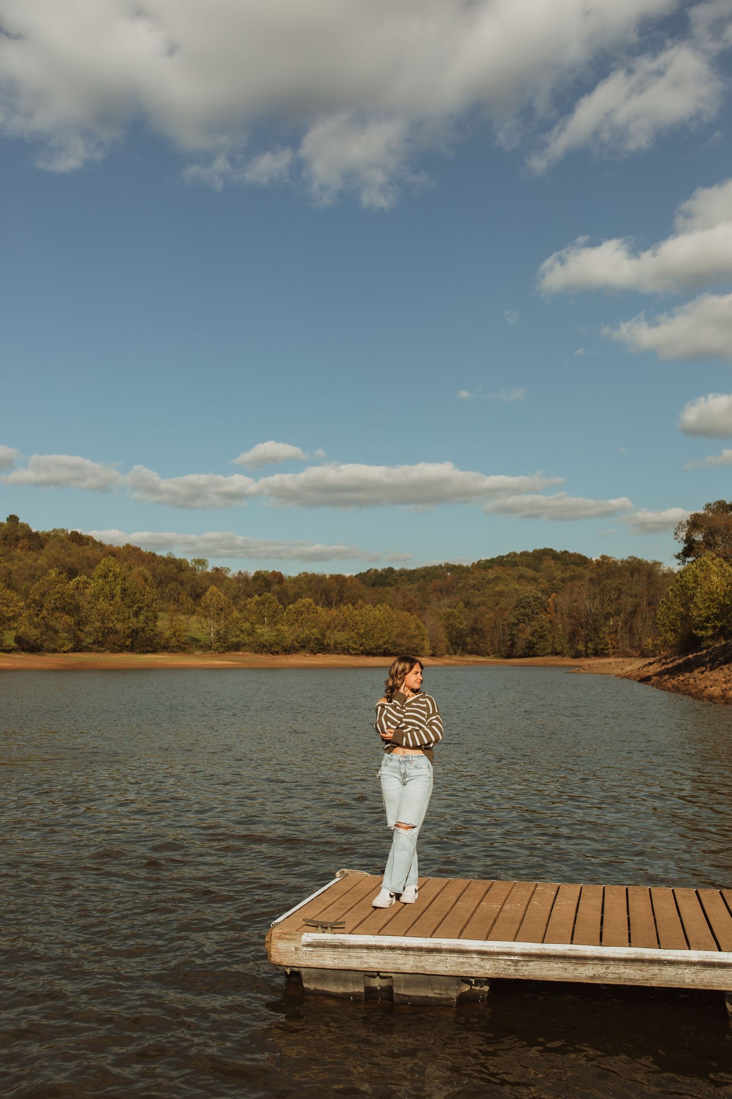 Figure in striped top stands on wooden dock overlooking serene lake surrounded by autumn hills.