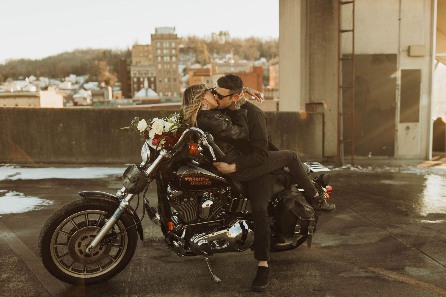 A couple poses with a motorcycle on a rooftop parking area with city buildings in the background.