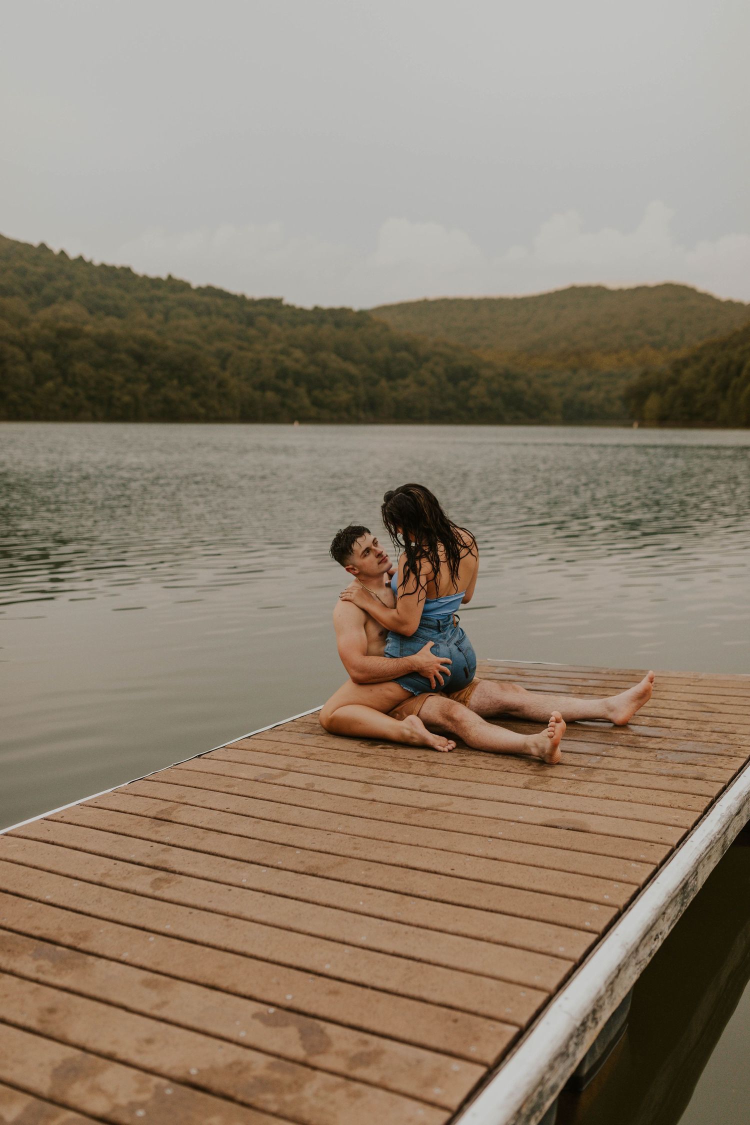 Couple sitting on wooden dock surrounded by mountains and lake.