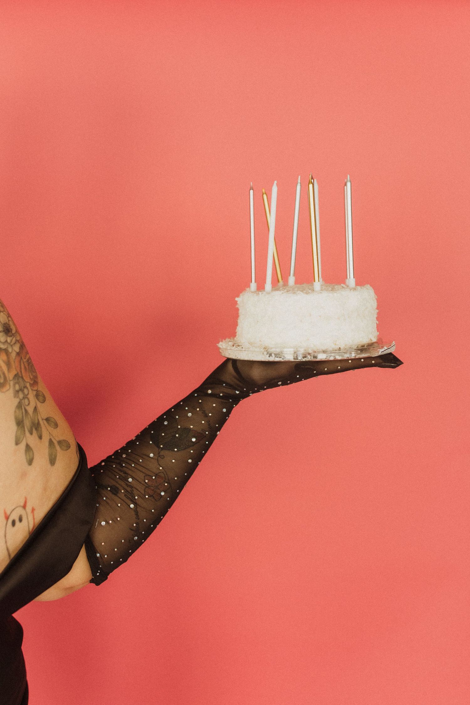 An arm in black lace gloves holding a small white cake with candles against a coral pink background. birthday photoshoot