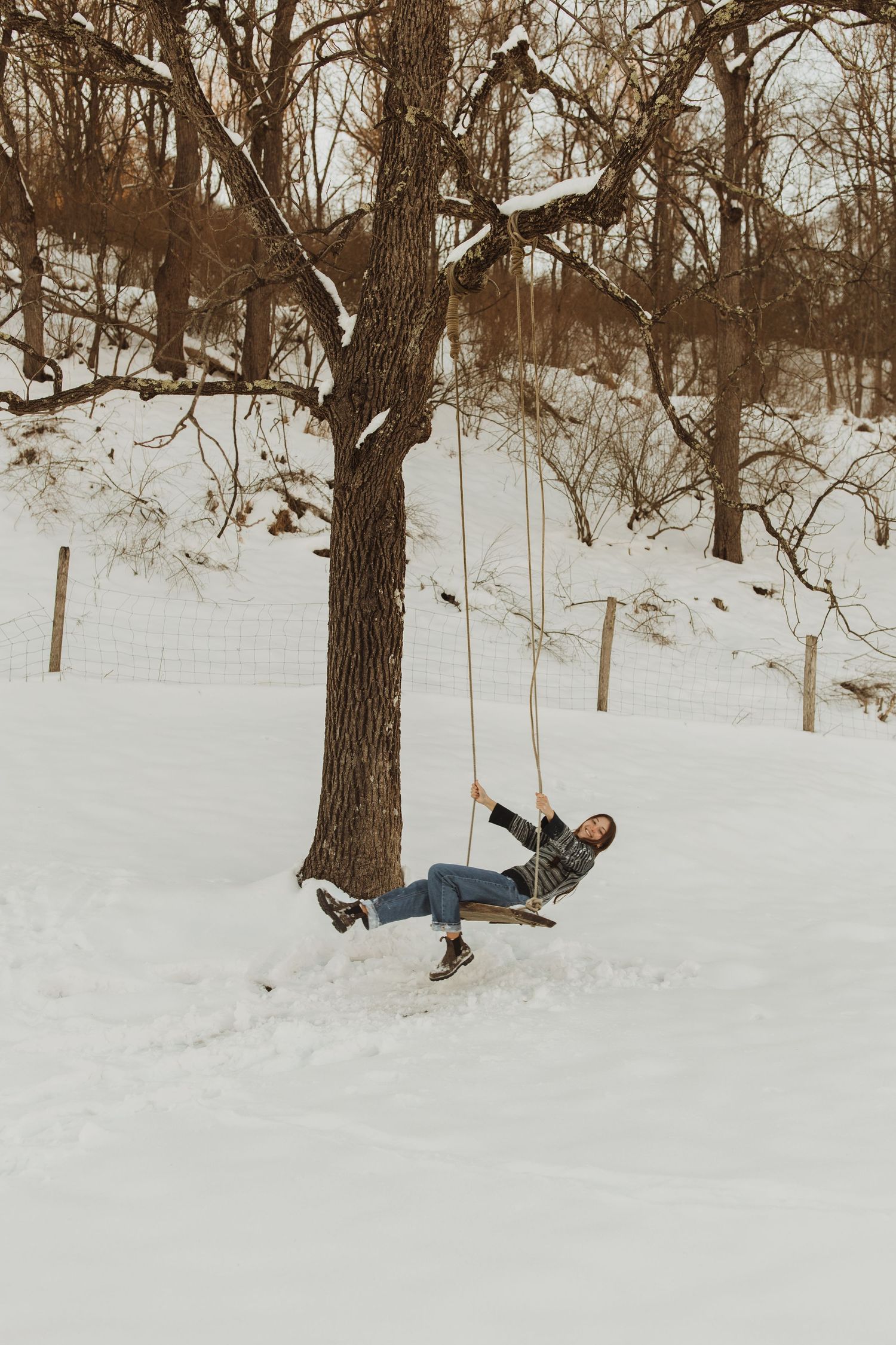 Person enjoying a playful swing from a tree branch in a snow-covered woodland area.