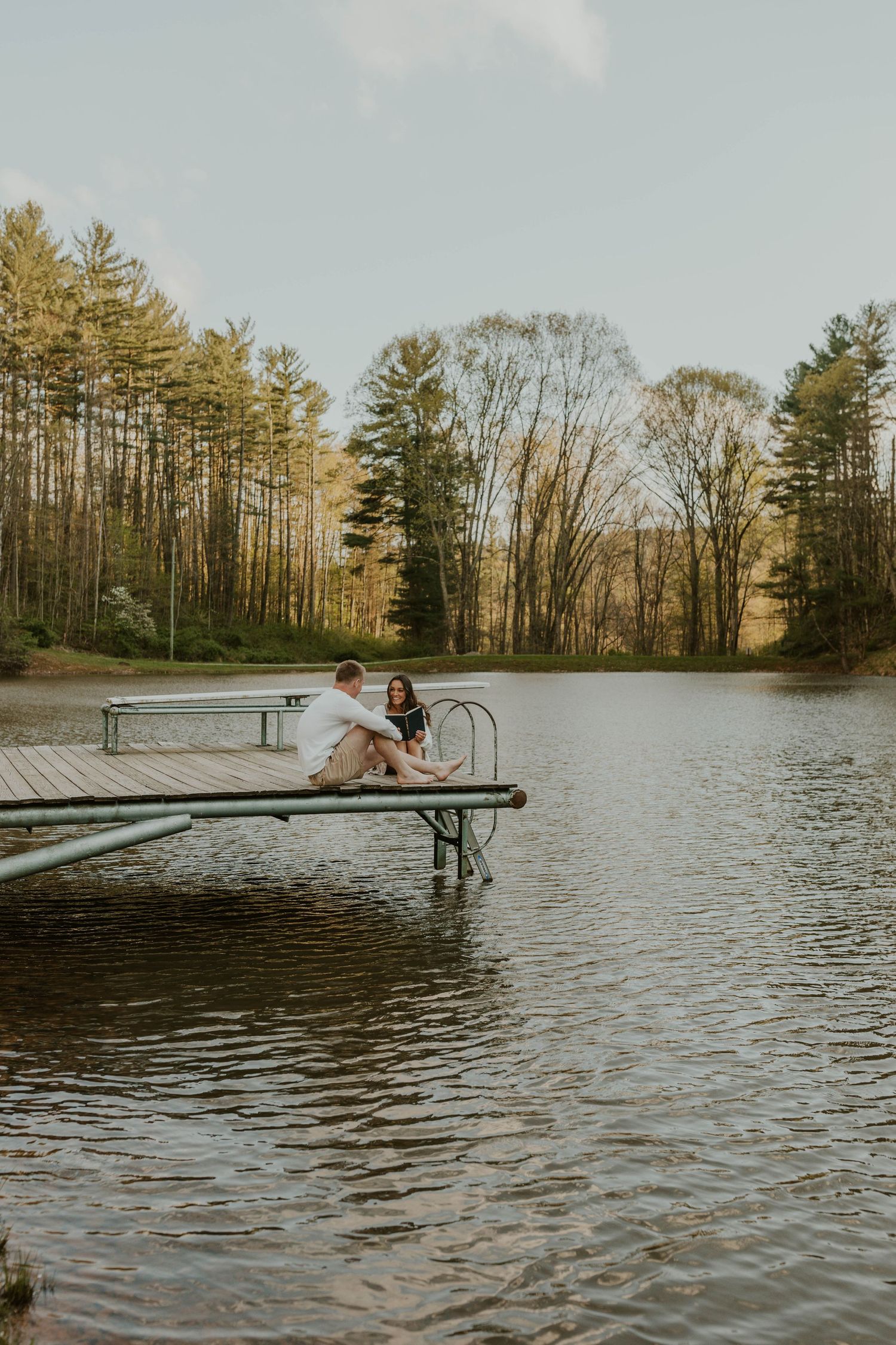 Two people sitting close together on lakeside dock surrounded by pine trees.