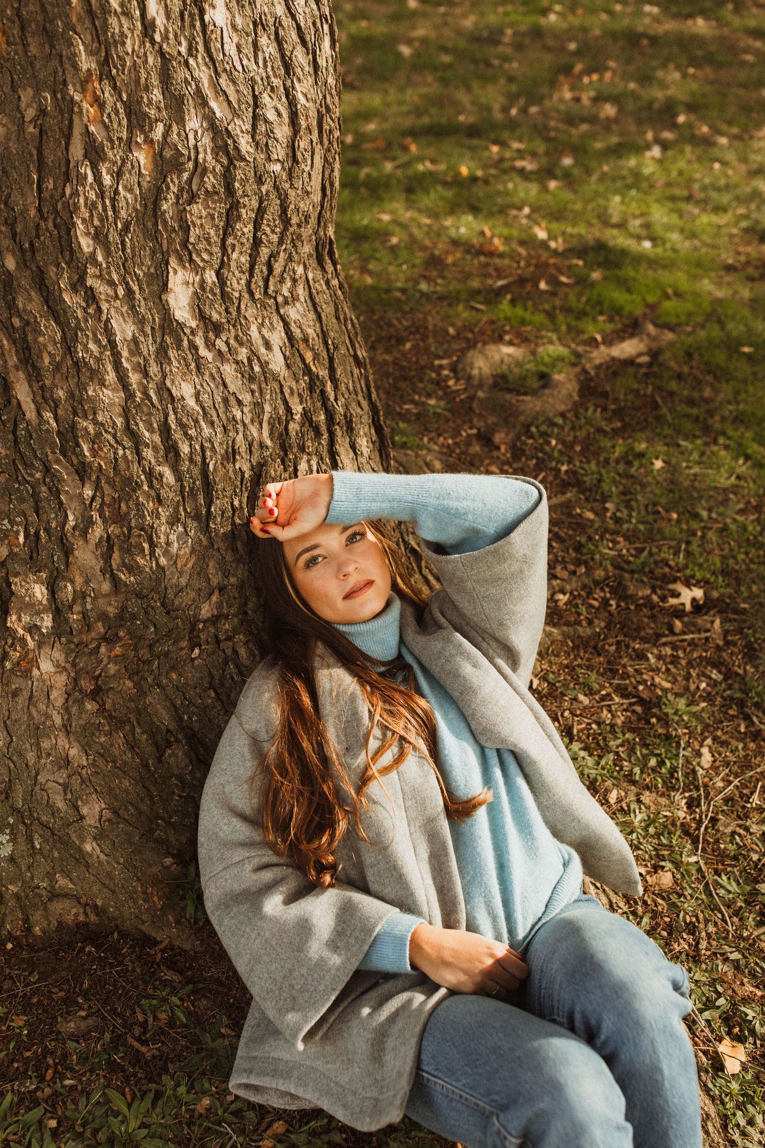 Person in blue jeans and gray coat relaxing against a large tree trunk.