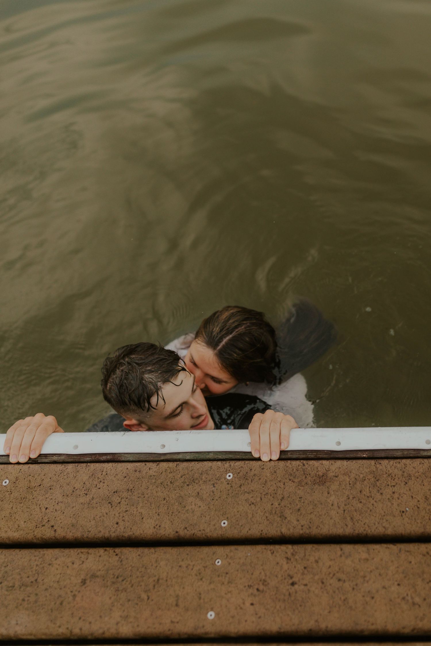 Two swimmers hanging onto a wooden dock while swimming in a dark lake.