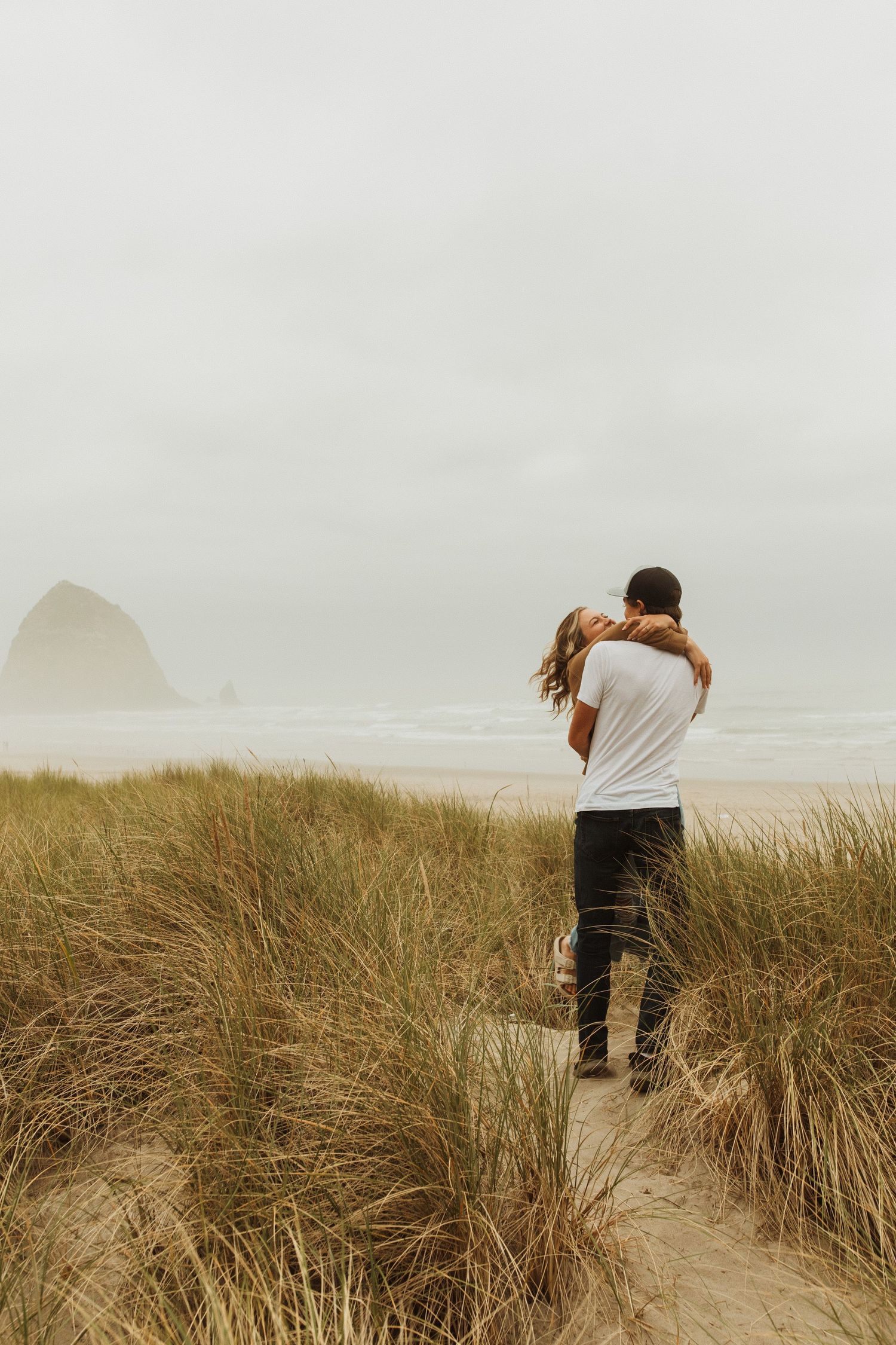 Foggy coastal scene with couple embracing on sand dune overlooking Haystack Rock.