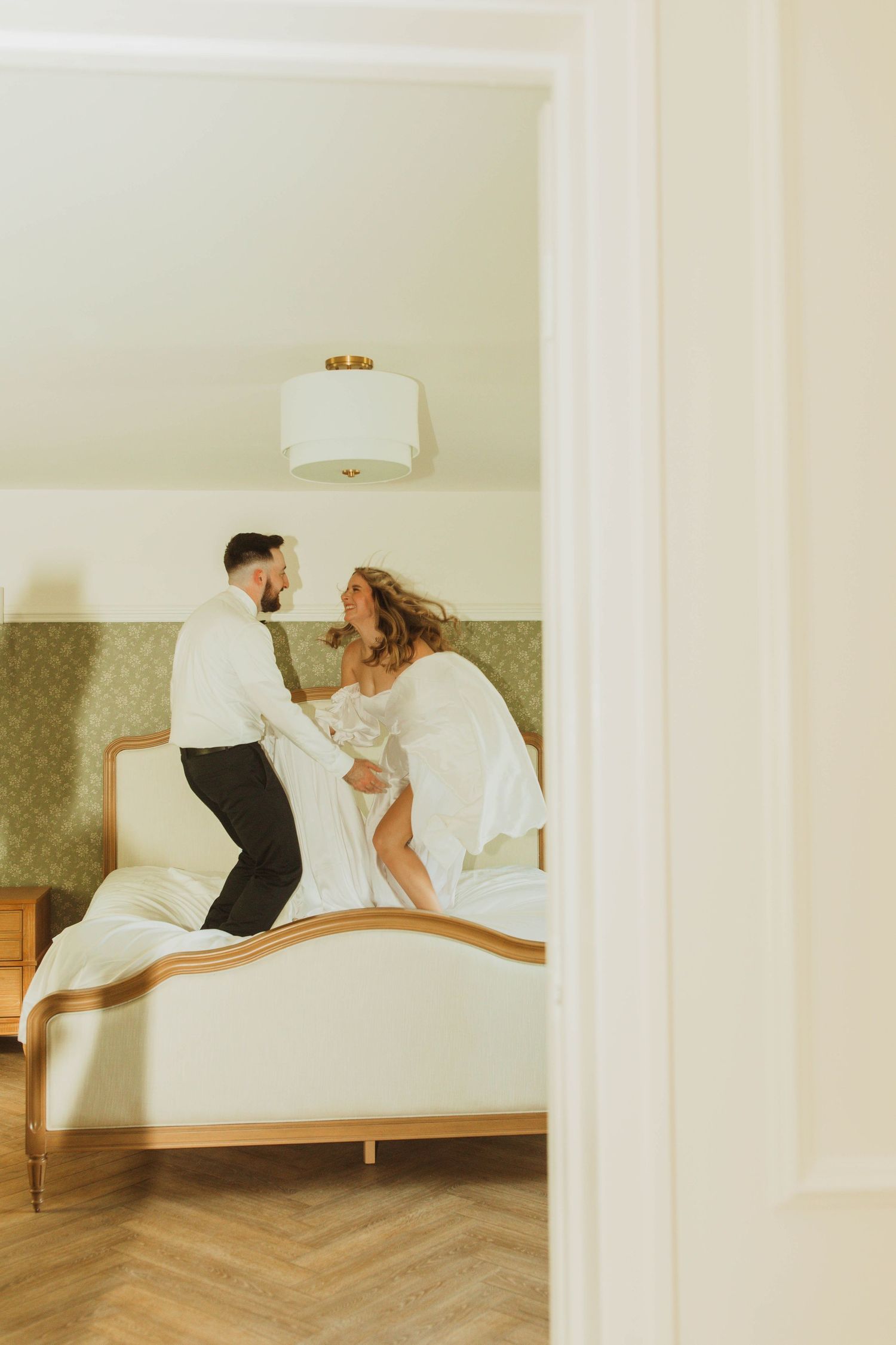 A playful wedding couple jumping on a bed in a softly lit room.