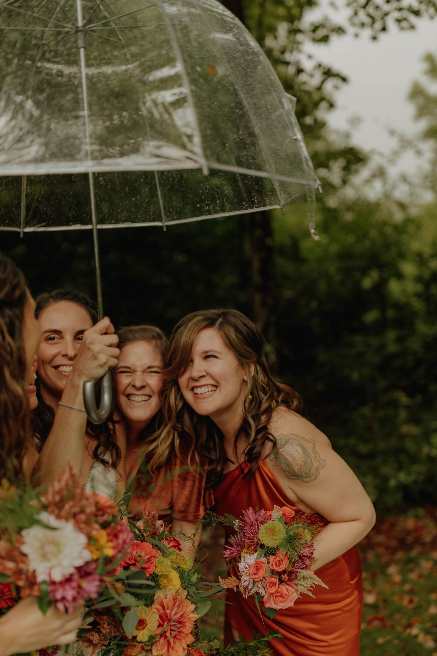 Joyful wedding party sharing laughter under a clear umbrella with colorful bouquets.