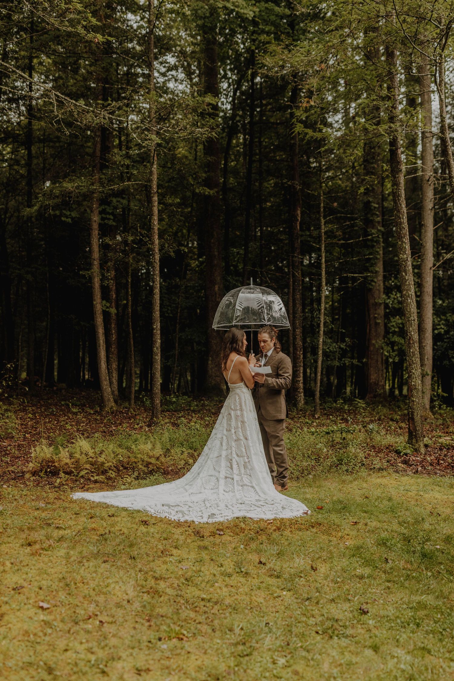 A wedding couple stands in a forest clearing with a long lace train dress.