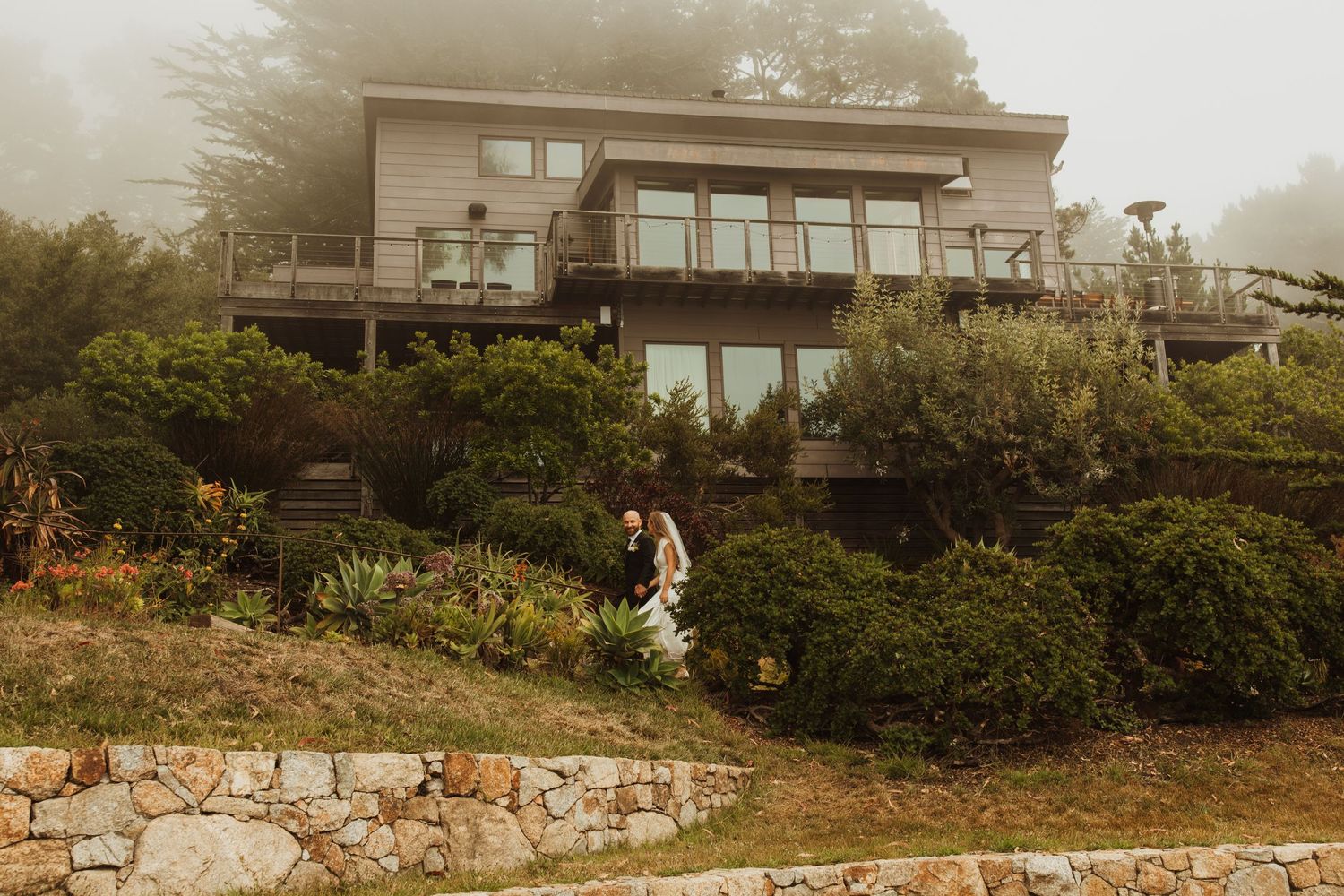 Modern hillside house with wedding couple walking on landscaped grounds.