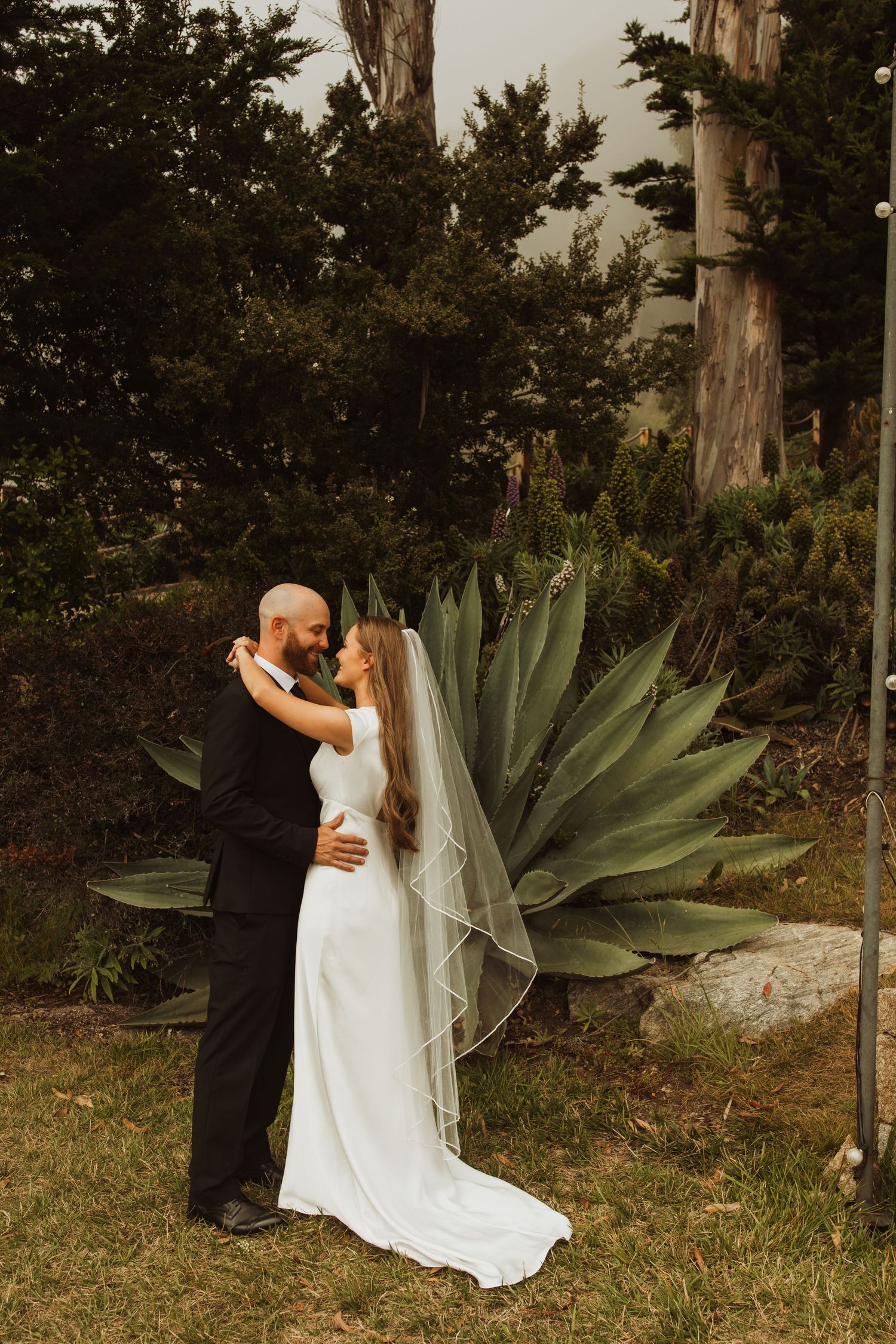 Romantic embrace in front of large agave plants and cypress trees during outdoor wedding.