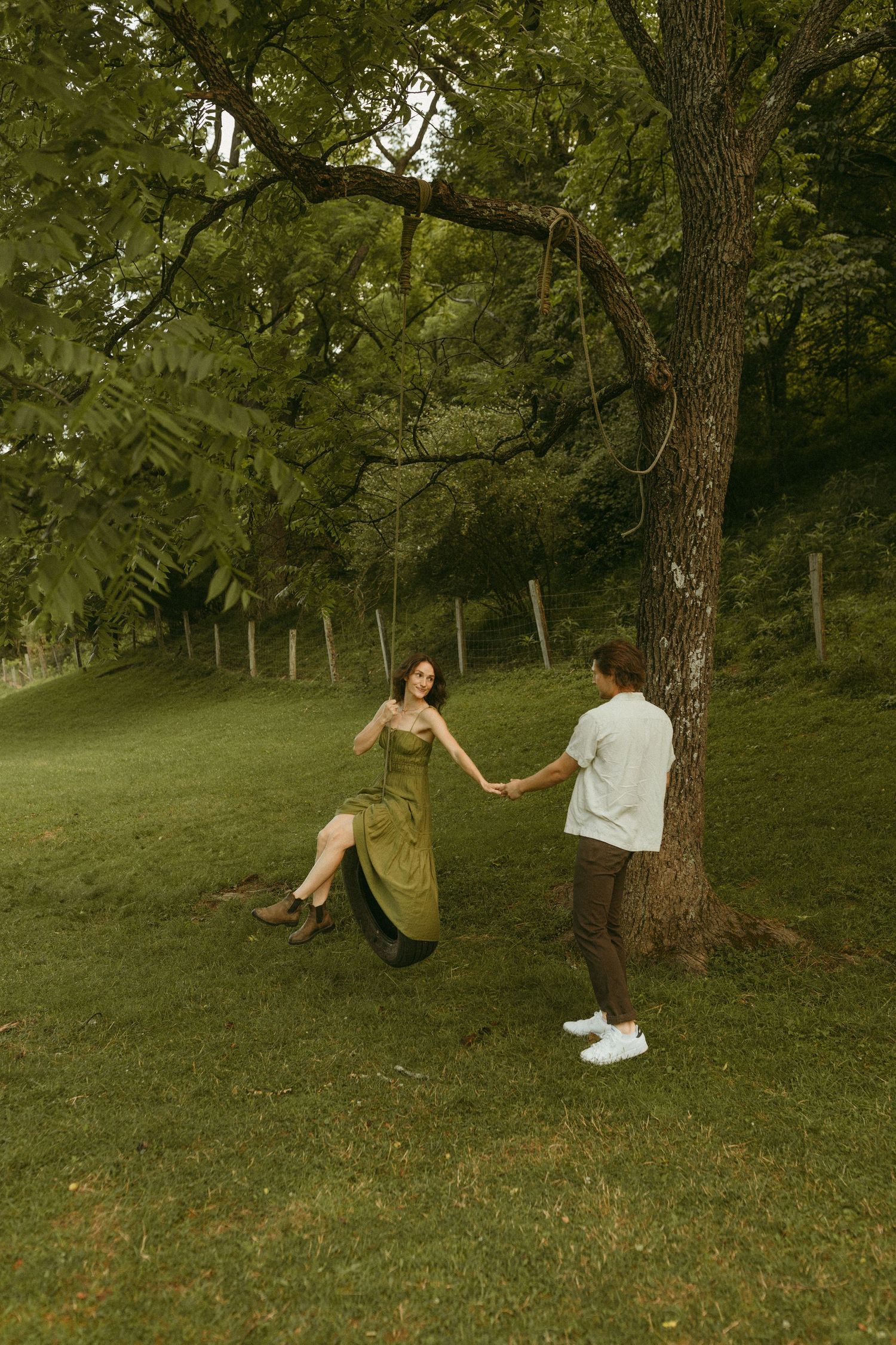 A couple playfully interacts near a tire swing on a grassy hillside surrounded by trees.