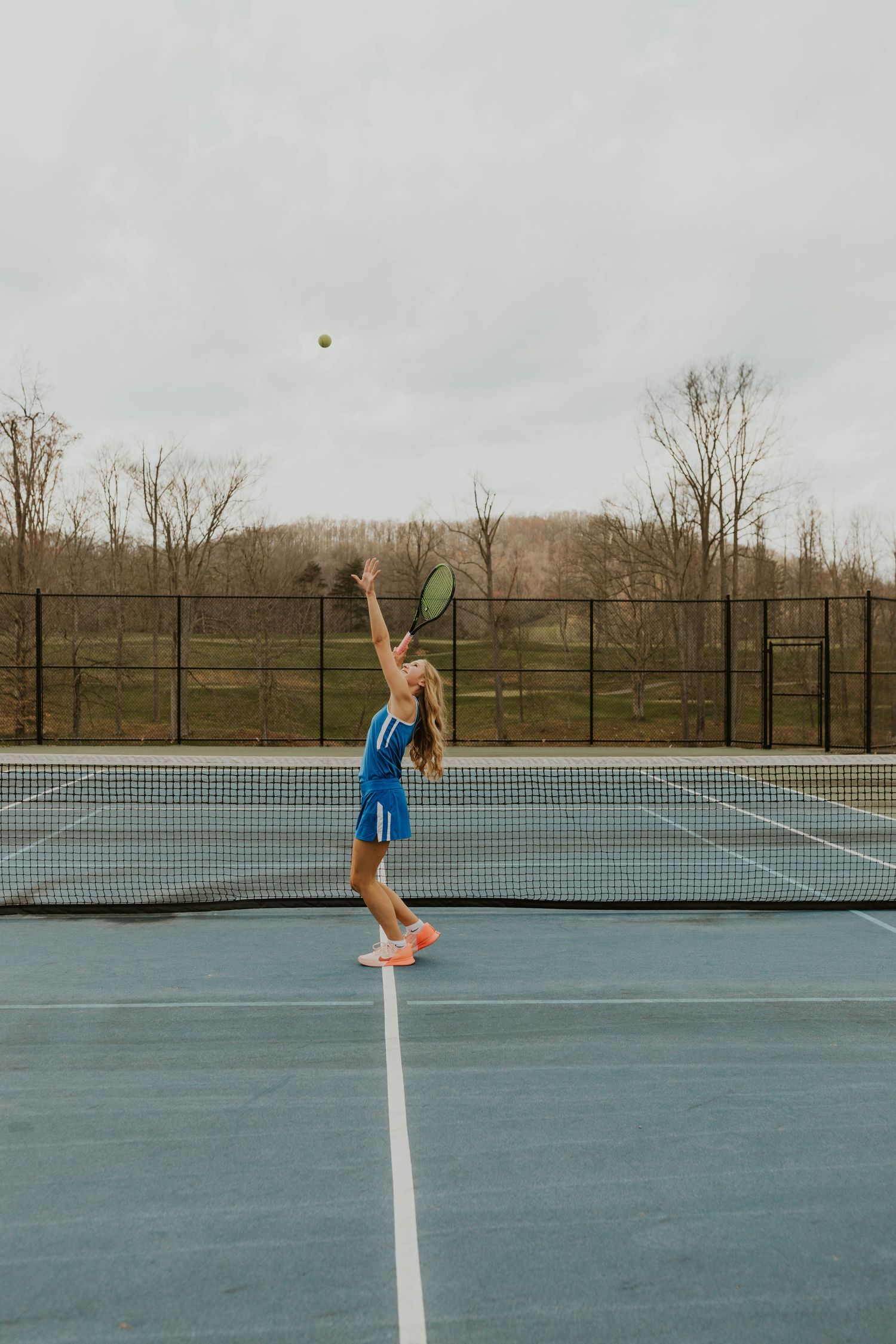 A tennis player serves the ball on an outdoor court surrounded by autumn trees.
