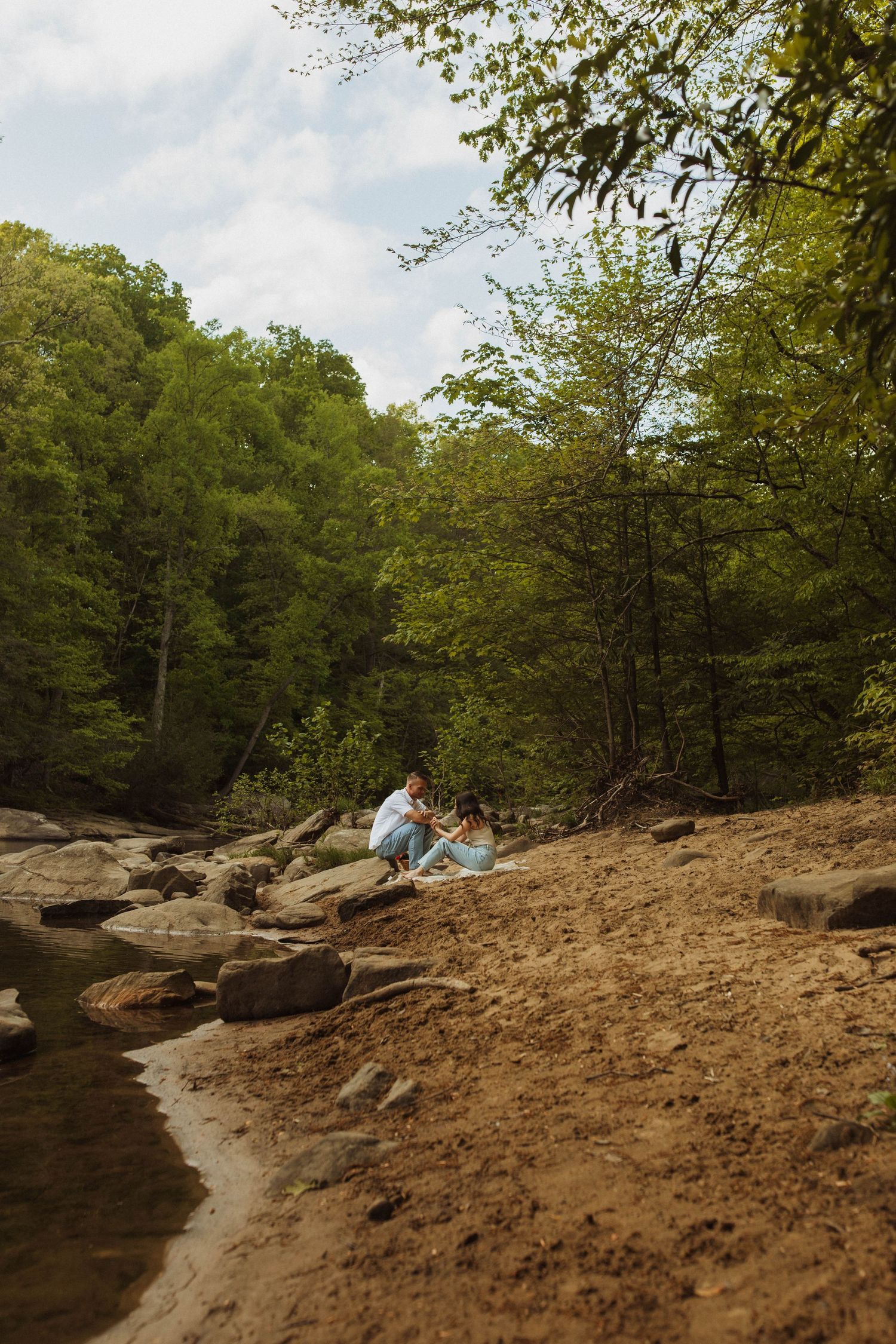 A couple relaxes on rocky terrain beside a serene creek in a wooded setting.