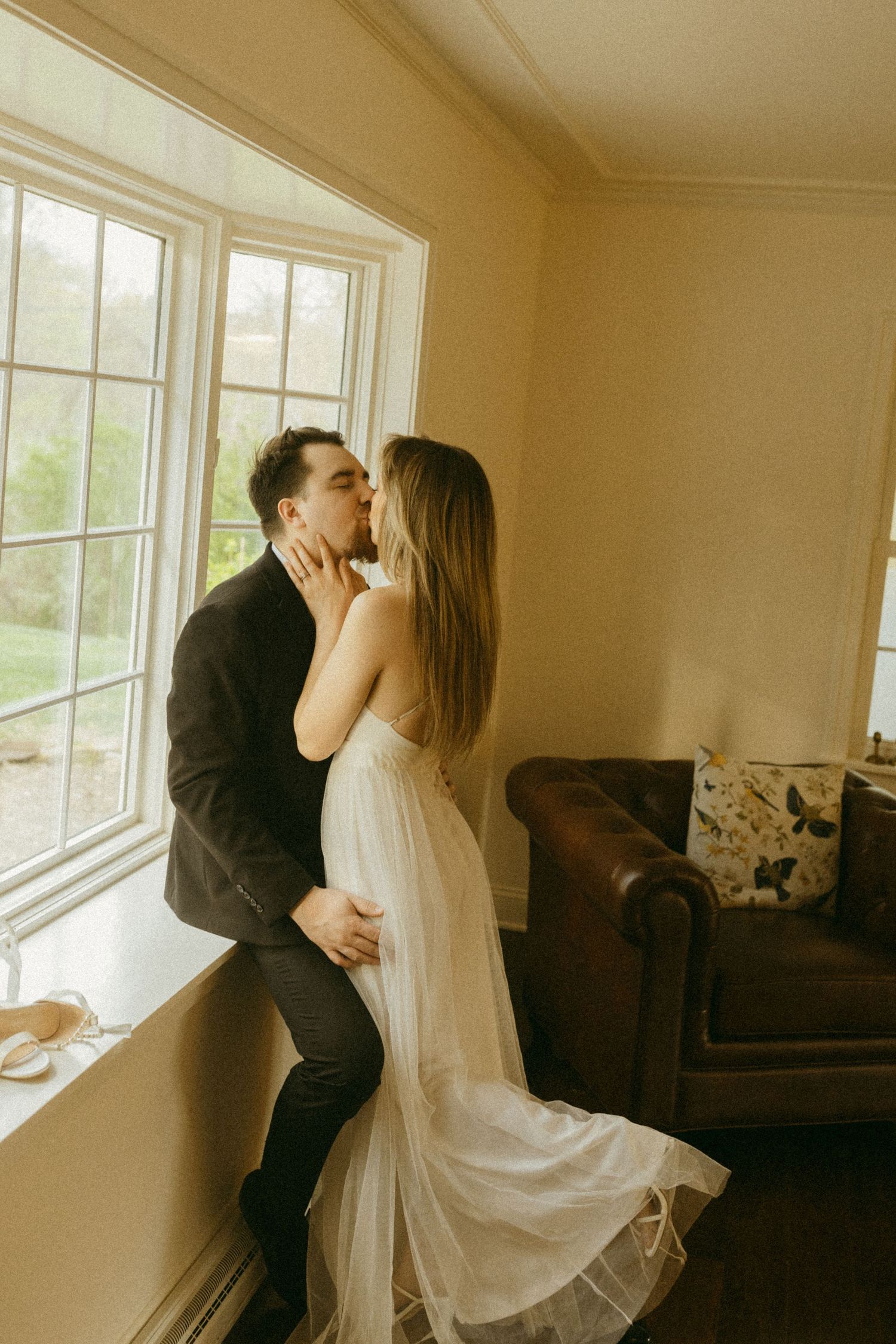 Romantic couple sharing an intimate kiss by a large window in a warmly lit room.