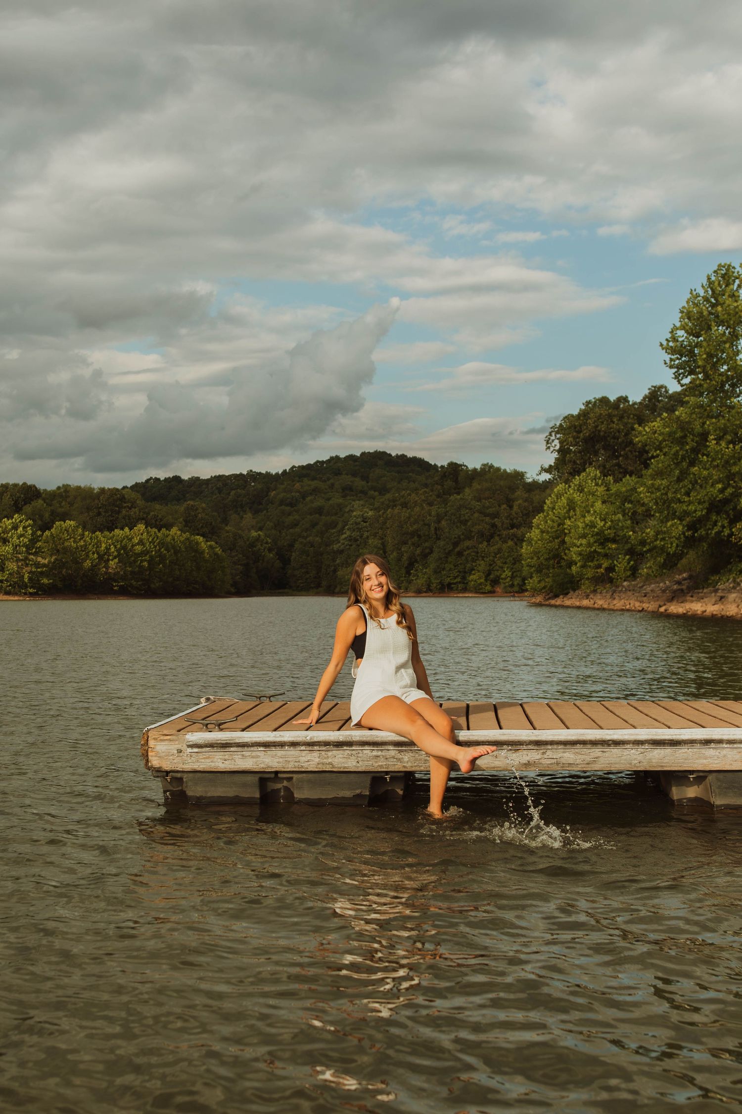 Person in white dress sits on wooden dock splashing feet in lake water at sunset.