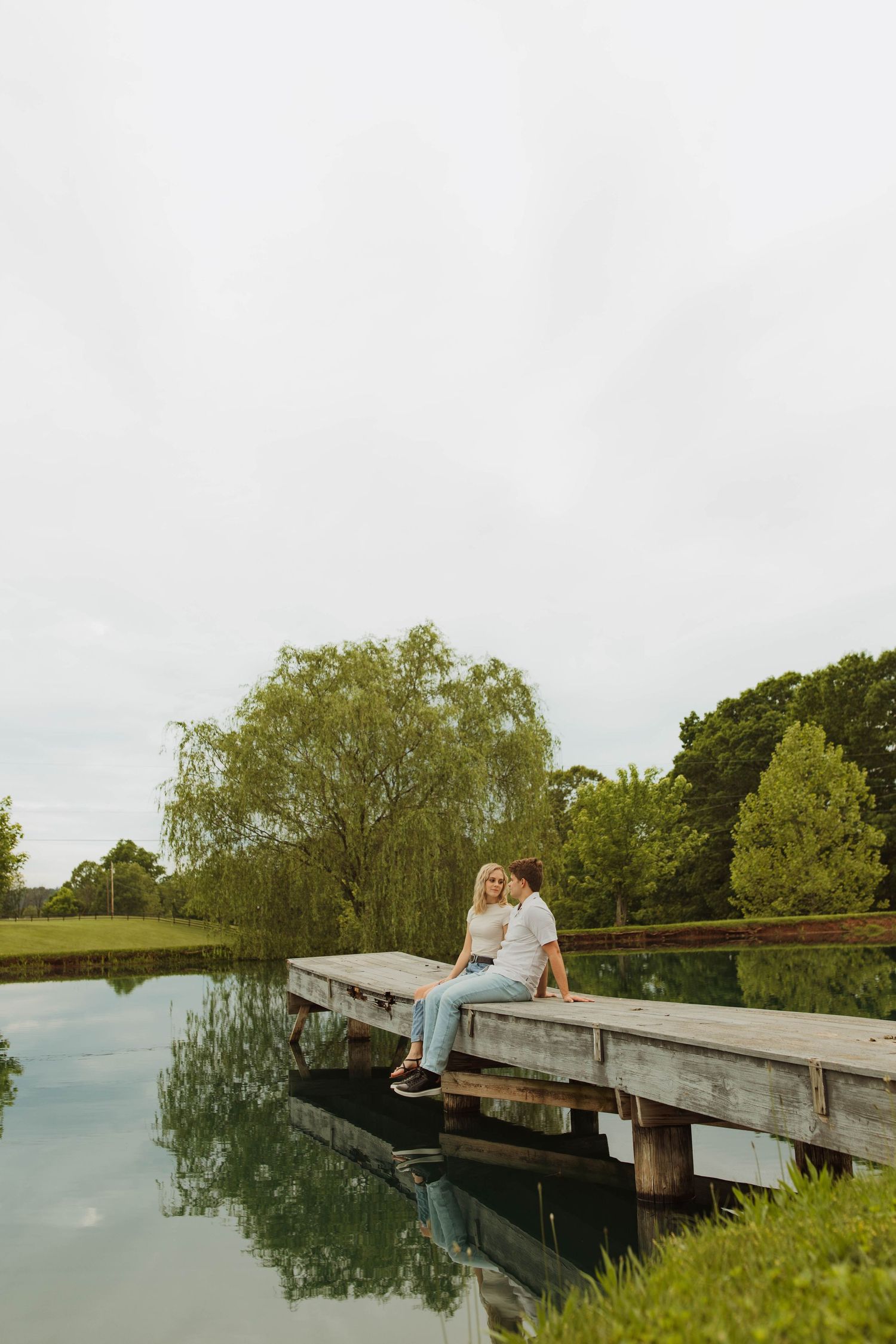 Couple sitting together on wooden dock over tranquil pond surrounded by trees.