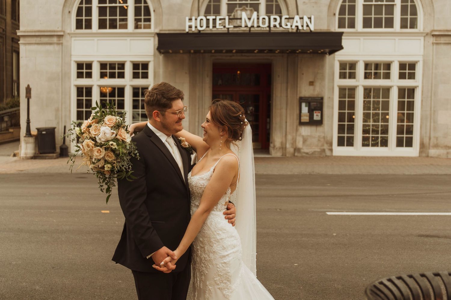 A wedding couple poses in front of Hotel Morgan holding a beautiful floral bouquet.