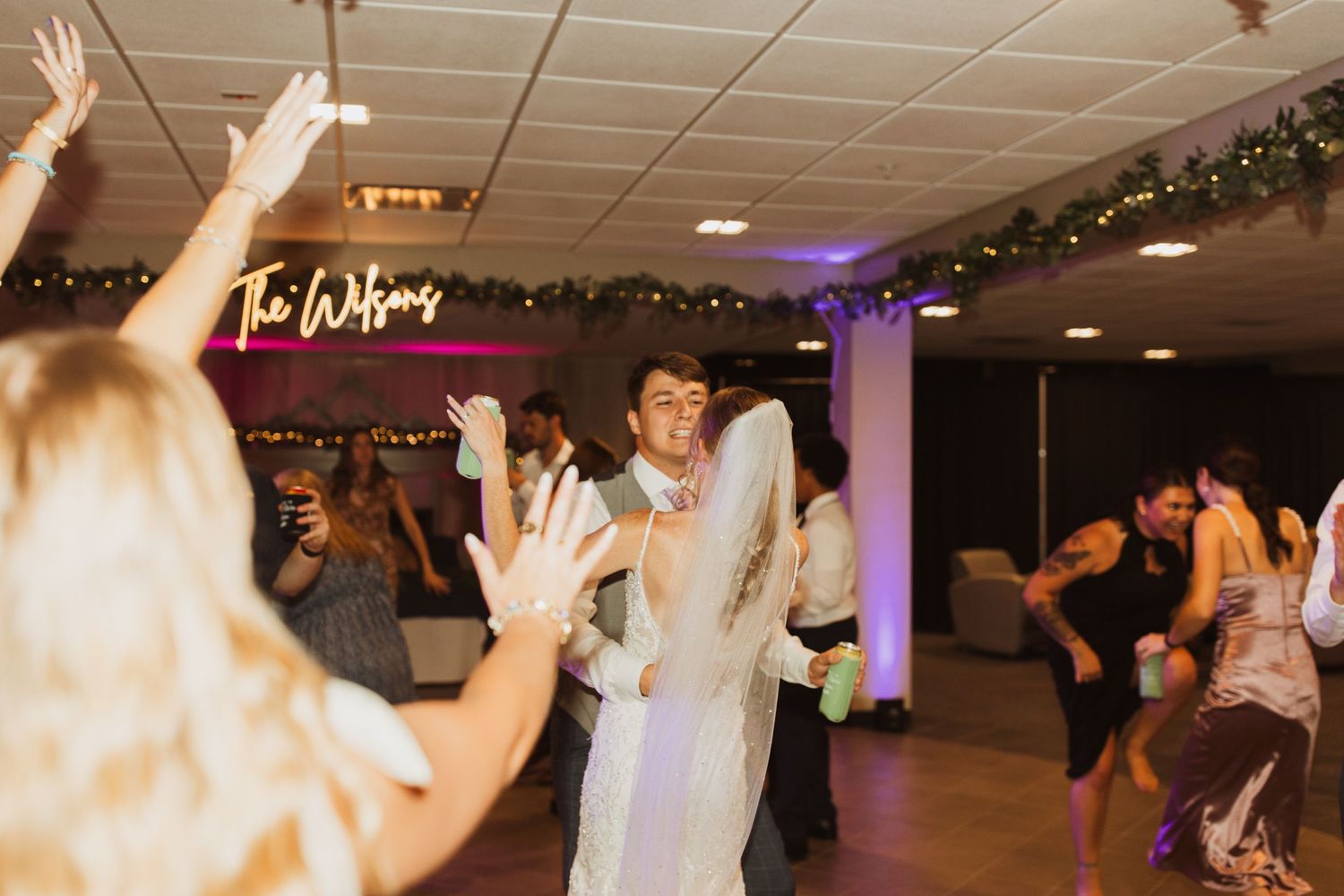 Wedding celebration in full swing with dancing and raised hands under purple lighting.