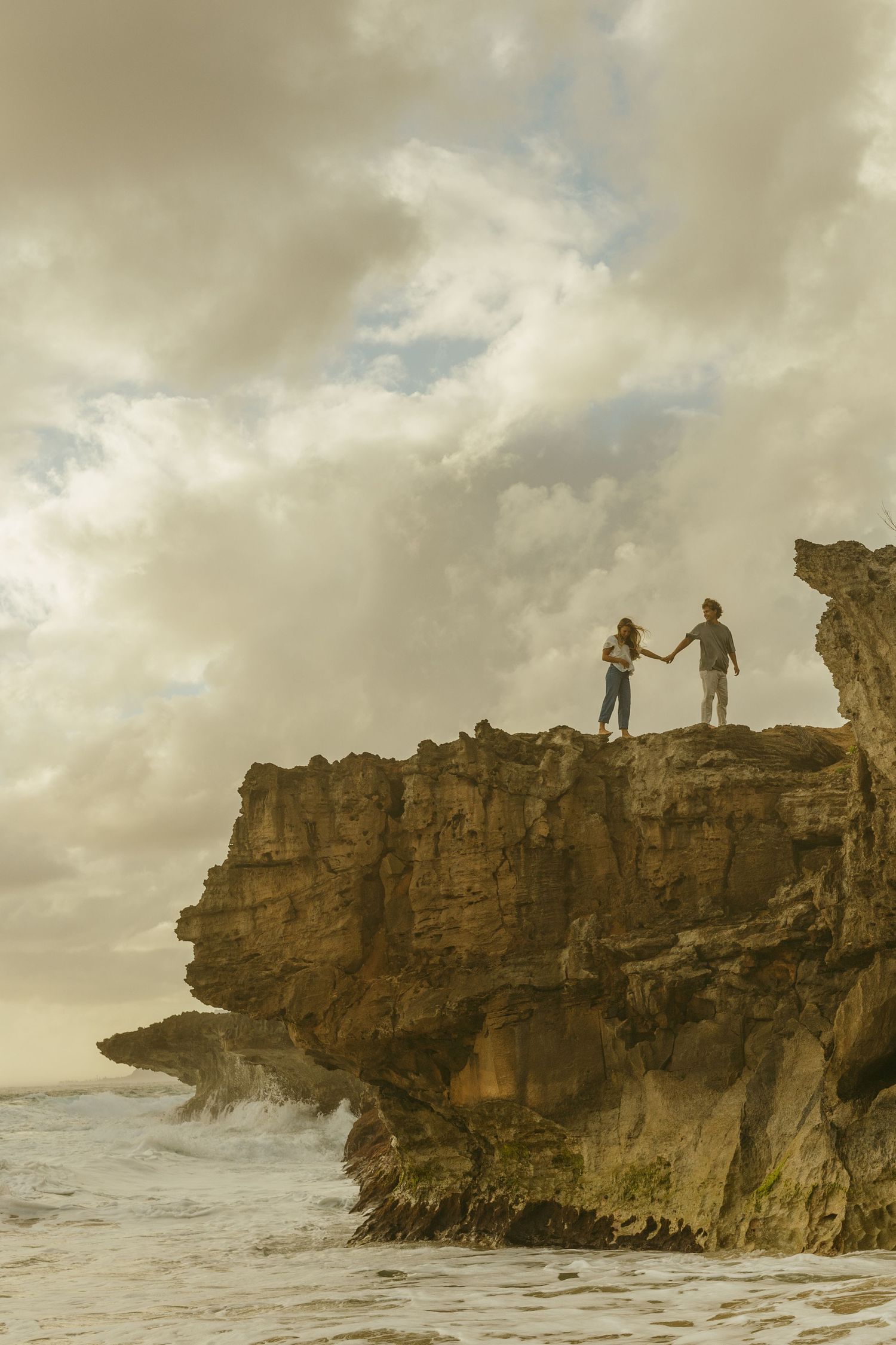 Two silhouettes standing on dramatic clifftop overlooking ocean waves at sunset.