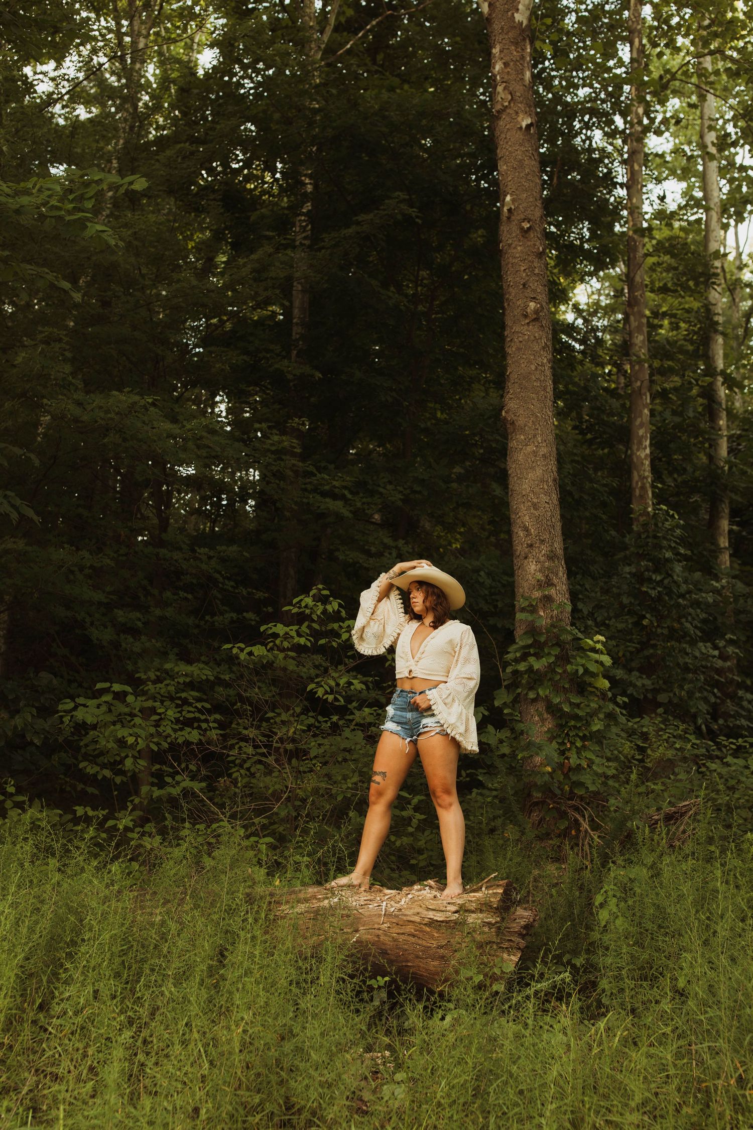Figure stands on log in forest wearing denim shorts and white top with cowboy hat.
